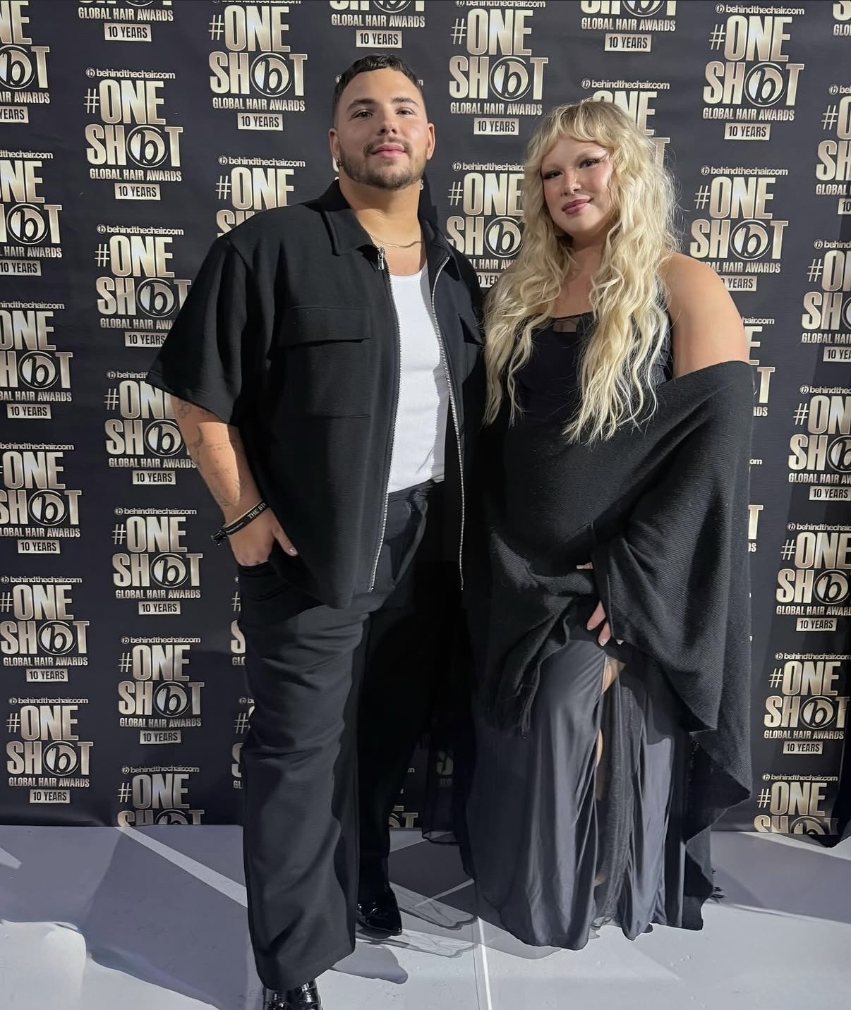 A man and a woman standing together in front of a backdrop with the logo for the Global Hair Awards and the hashtag #OneShot, celebrating 10 years.