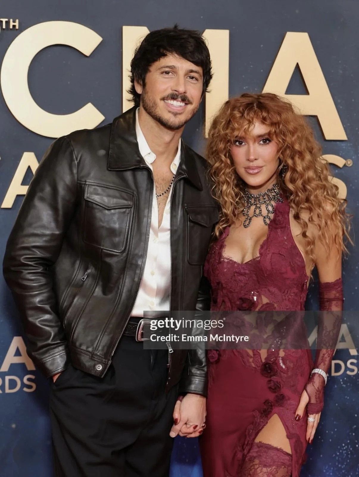 A man and woman holding hands at an awards event, standing in front of a blue background with large gold letters. CMA Awards