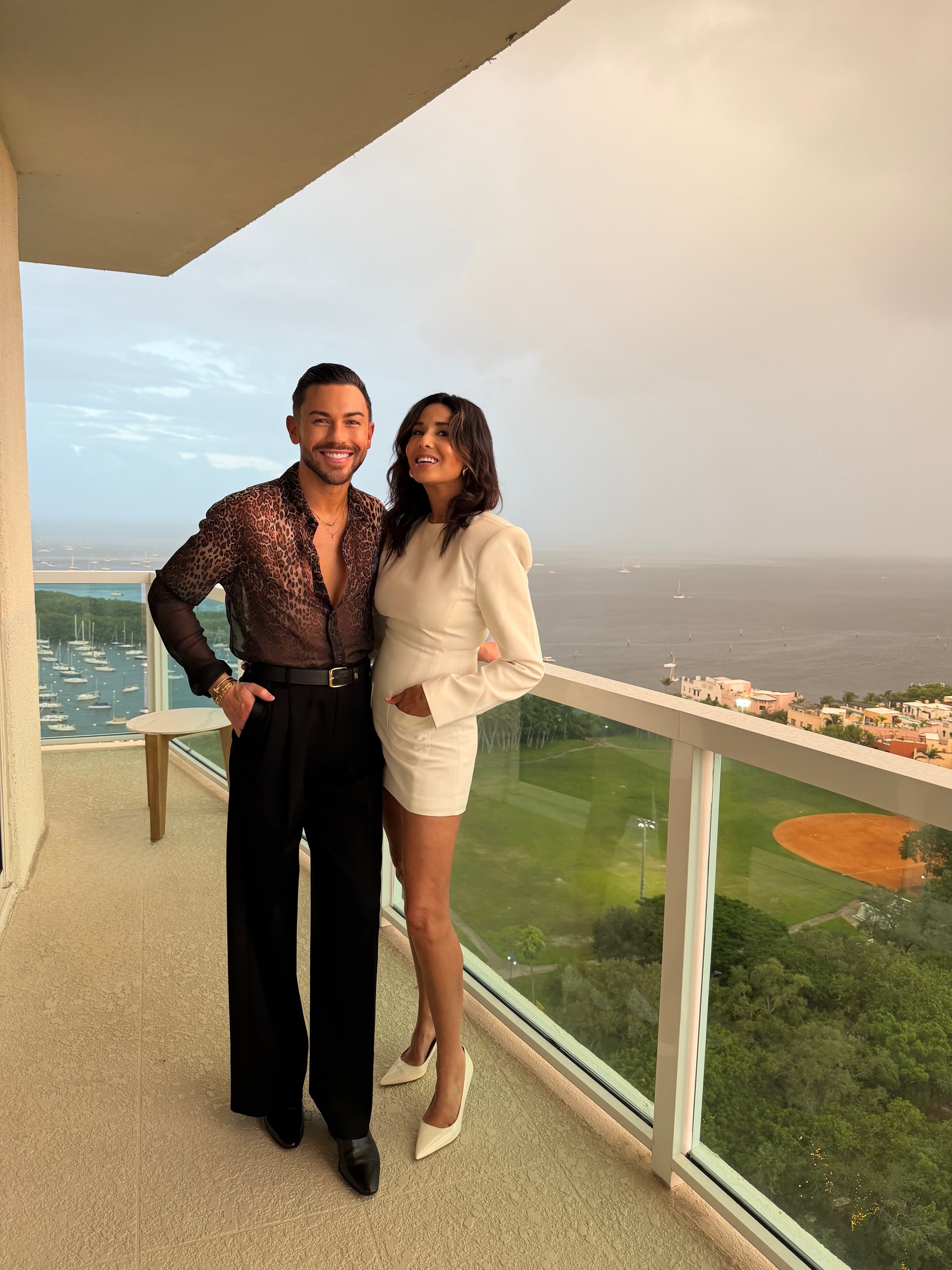 A man and woman standing on a balcony with a view of the ocean, boats, and greenery in the background; they are smiling and dressed in stylish clothing.