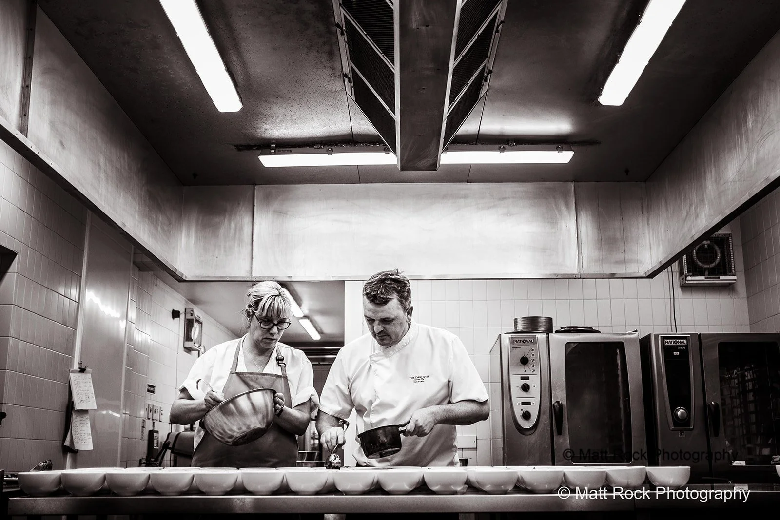 Black & White photo of a Commercial kitchen shoot in Kent of a chef and assistant making up the meals. 