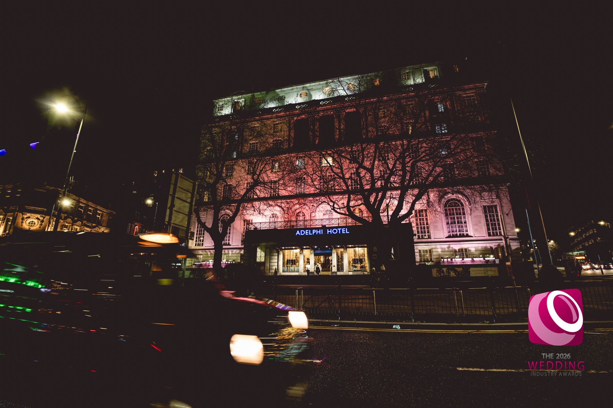 Night time photo of the outside of The Adelphi Hotel, Liverpool with a black taxi driving passed