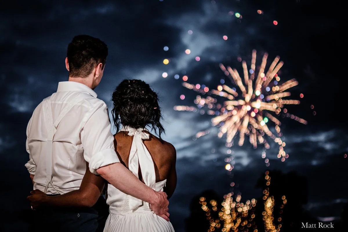Colour photo of fireworks in the distance and looking at the bride and groom watching them explode into lots of sparkly colours.