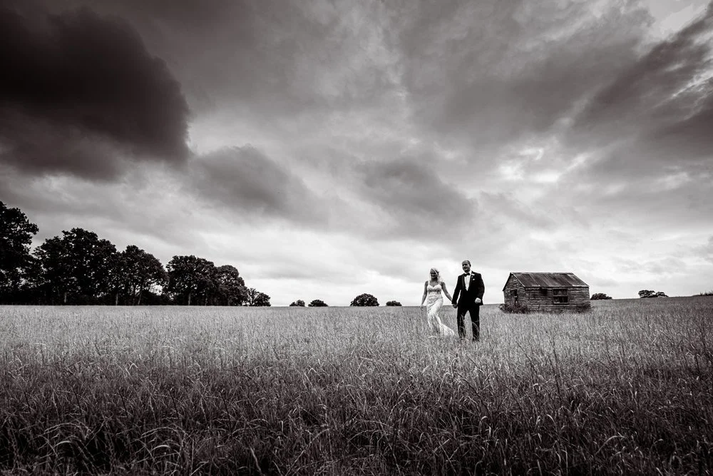 Black & White photo of the bride and groom walking in a field with a wooden house in the back ground. Wedding Photographer in Kent