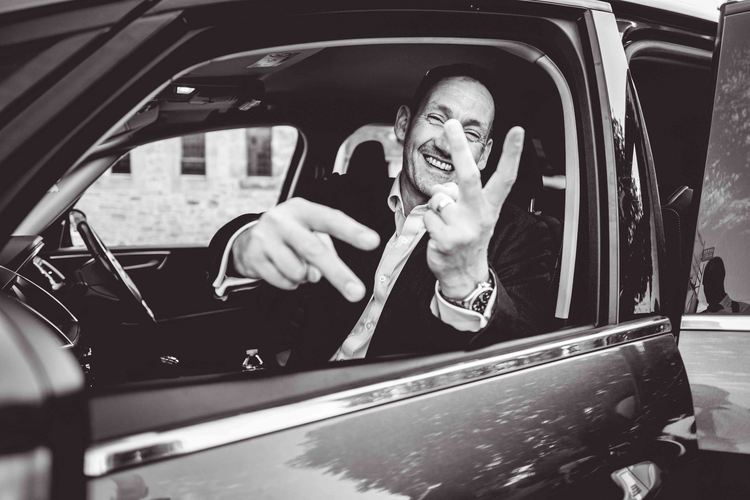 Black and white photo of one of the guests in a car sticking his fingers up to me - the photographer - during a funny exchange at Hever Castle, Kent, UK.