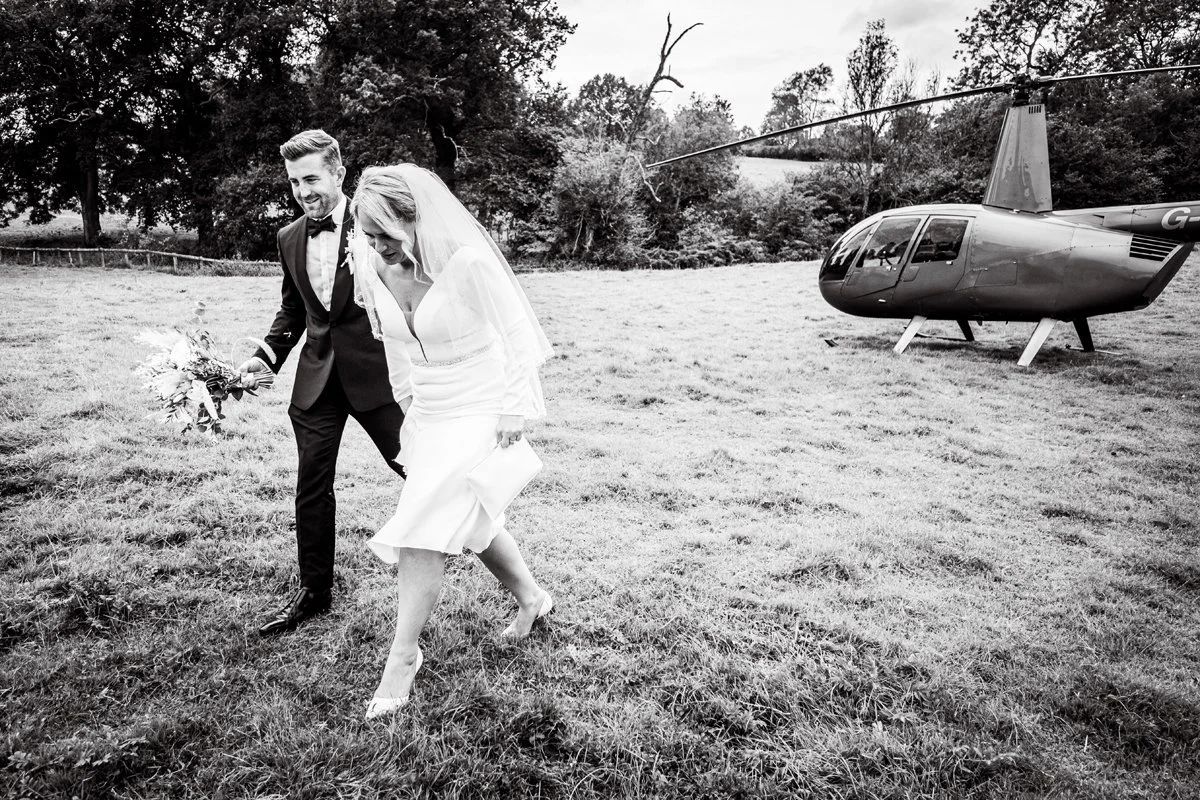 Black and white photo of happy bride and groom walking in field away from their helicopter after just flying in and arriving at their wedding venue at The Oak Barn Frame Farm