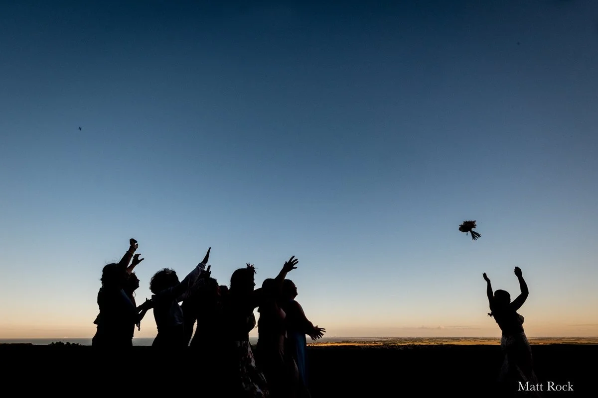 Silhouette of the Bride on the terrace with the sun setting in the background whilst throwing her bouquet to her friends at Lympne Castle, Kent - UK