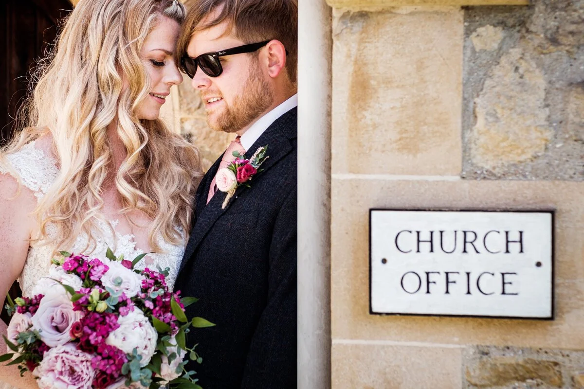 Bride and Groom portraits outside their Church - Groom is wearing sunglsses and a nice suit and Bride is in her wedding dress and holding flowers. Wedding photographer Kent