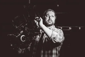 White man wearing checked shirt holding a video camera during a shoot - London Portrait Photographer Matt Rock