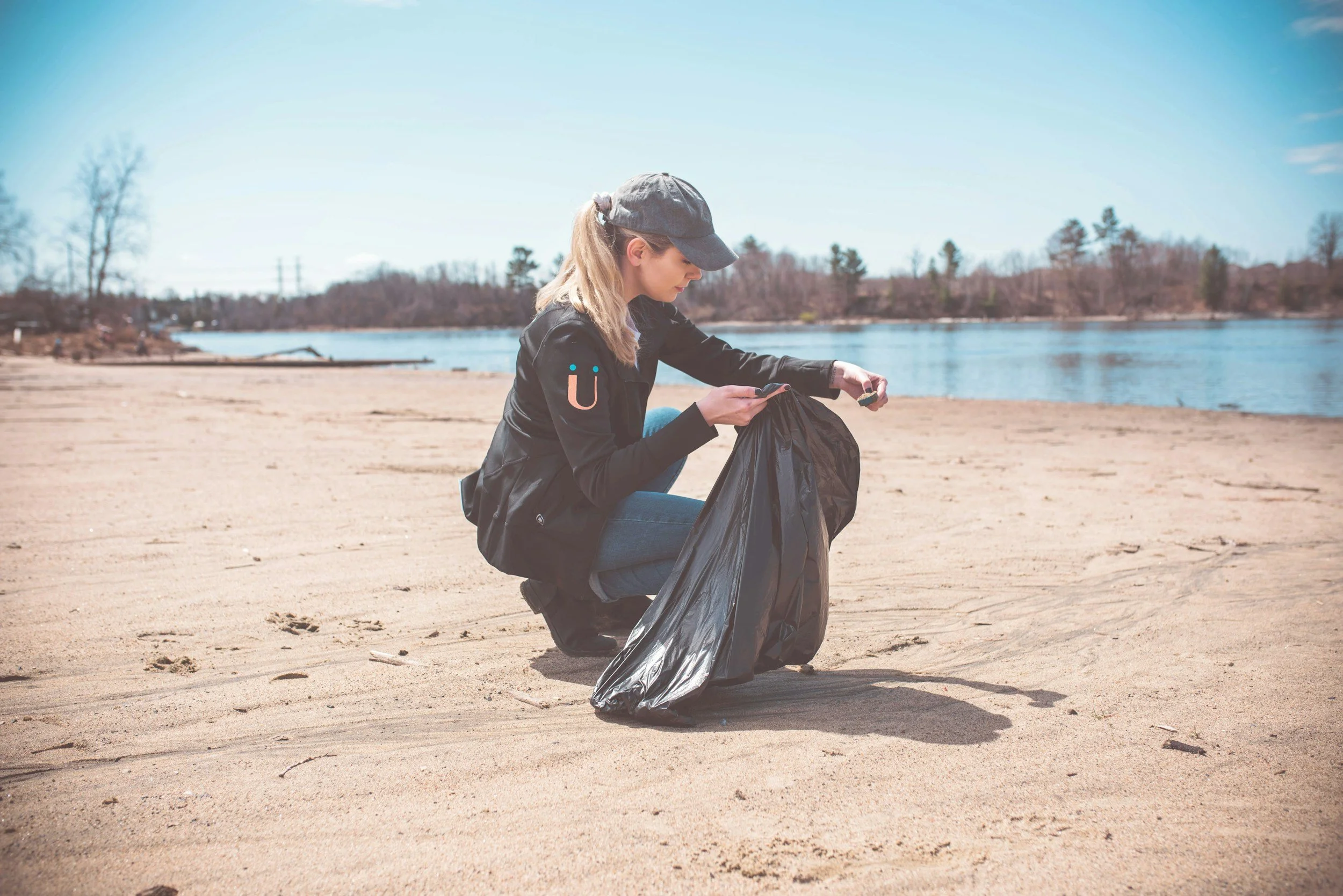 A woman cleaning up trash on a sandy beach by a river, holding a black trash bag and using a small tool to pick up debris.
