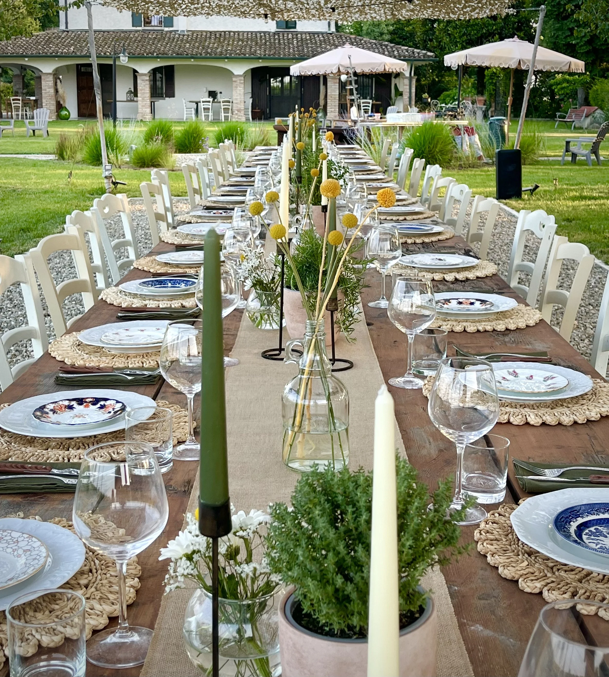 Tavolo all'aperto decorato in stile rustico con fiori, candele e stoviglie vintage, pronto per un evento in giardino con sedie bianche, ombrelloni e giardino curato intorno.