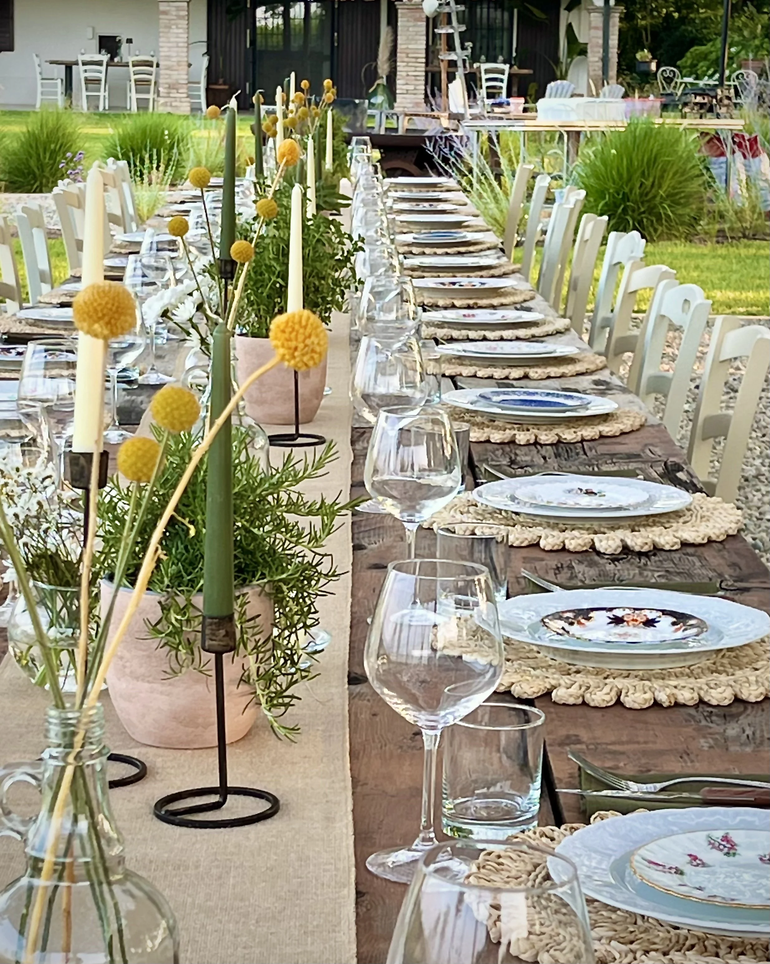 Tavola all'esterno con piatti, bicchieri e candelabri, decorata con fiori gialli e piatti vintage, in un ambiente verde, curato e tranquillo.