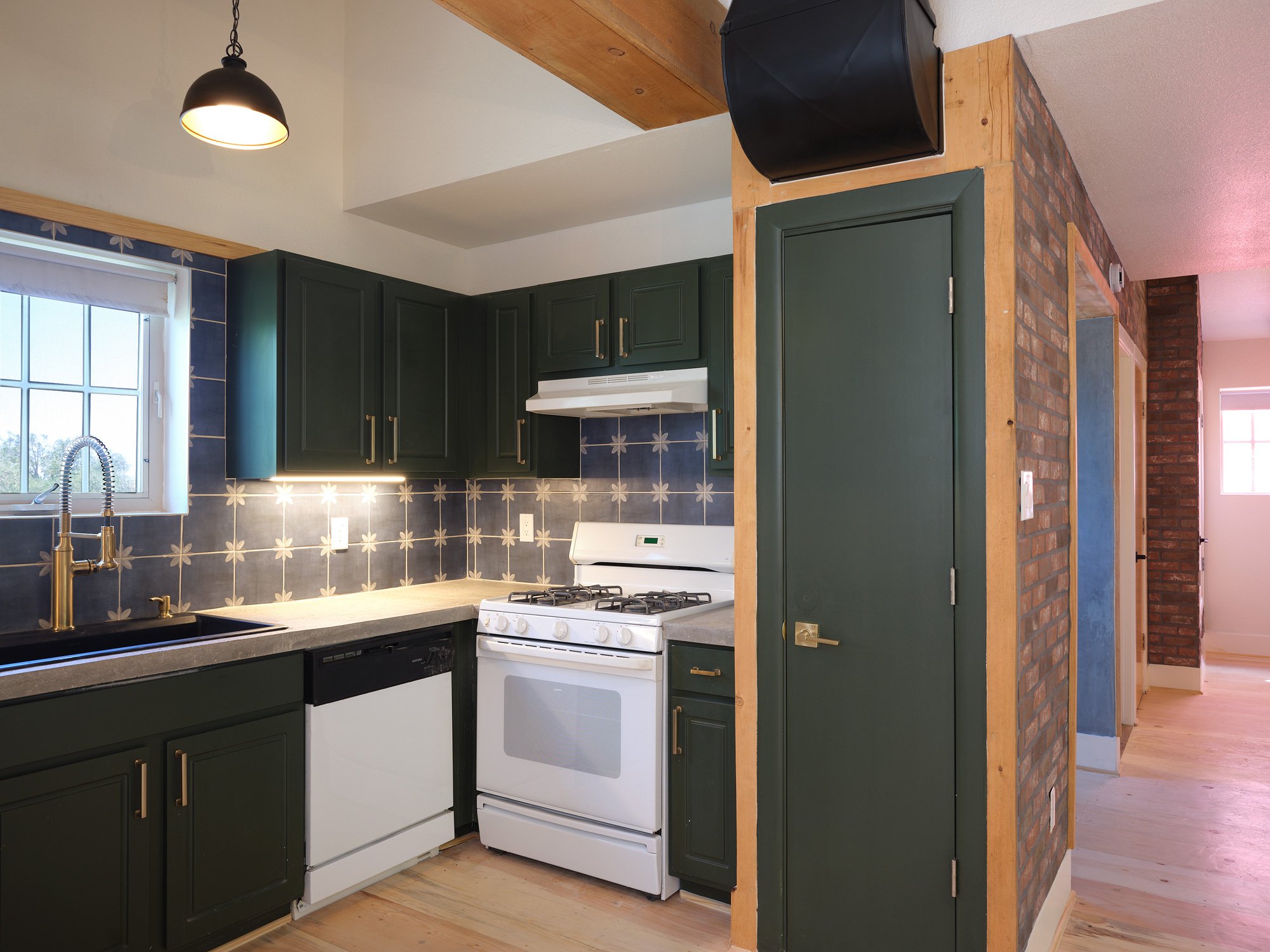 Kitchen interior with dark green cabinets, a white stove, grey countertop, a window with a white frame, and a black hanging light fixture.
