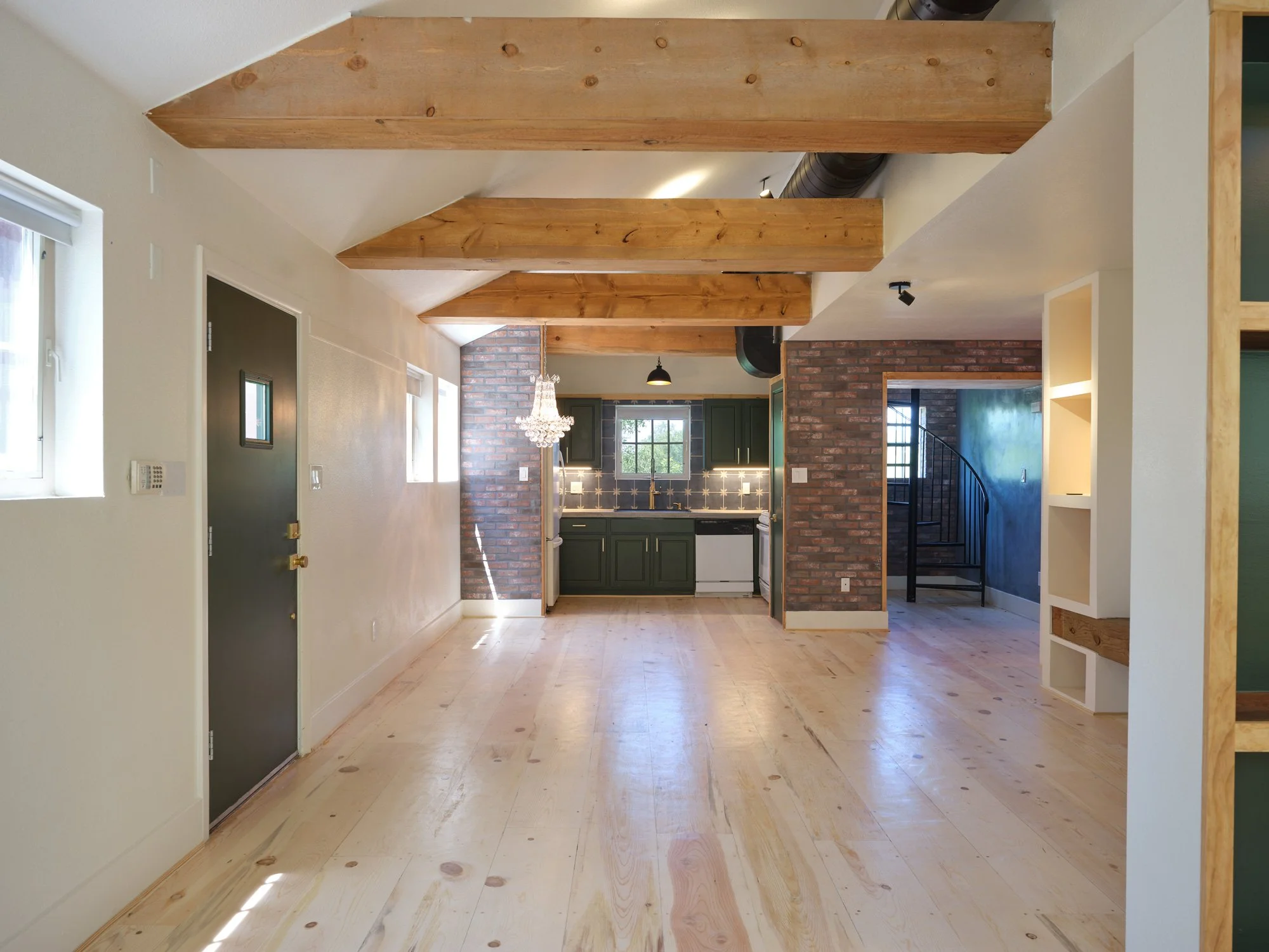 Interior view of a modern living space with wooden beams on the ceiling, light-colored wooden floors, a kitchen area with green cabinets and brick wall accents, and a spiral staircase in the background.