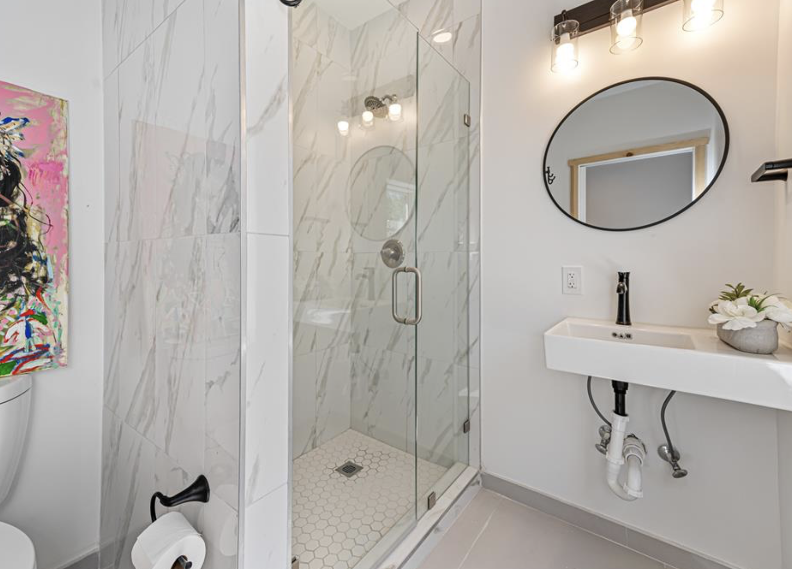 Modern bathroom with a glass-enclosed shower featuring marble walls, a white rectangular sink with black fixtures, a round mirror, a flush-mounted ceiling light fixture, a colorful abstract painting on the wall, a small potted plant on the sink, and a gray toilet paper holder.