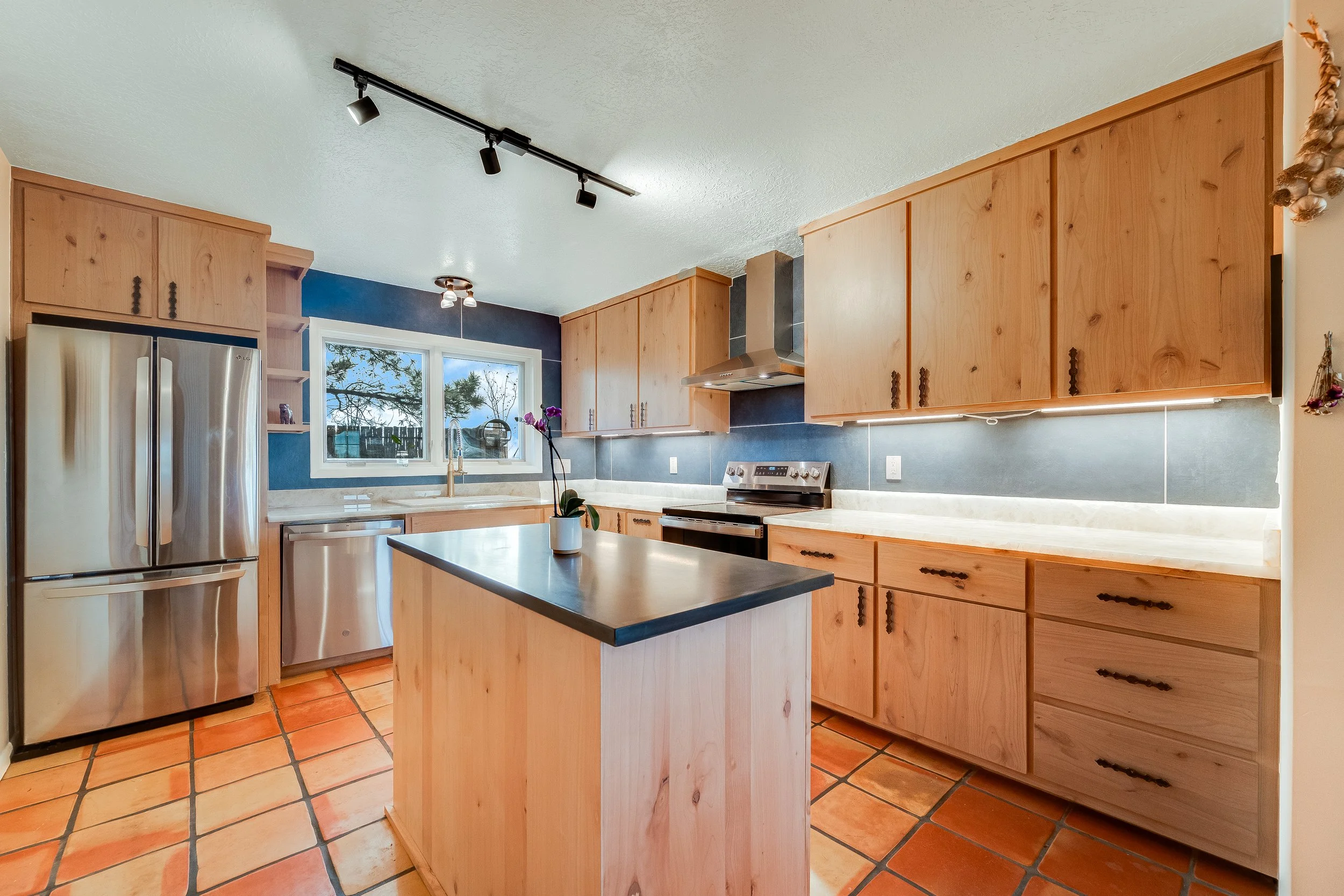 Kitchen with wooden cabinets, stainless steel appliances, a small island, orange tiled floor, window above the sink, and a potted orchid on the island.