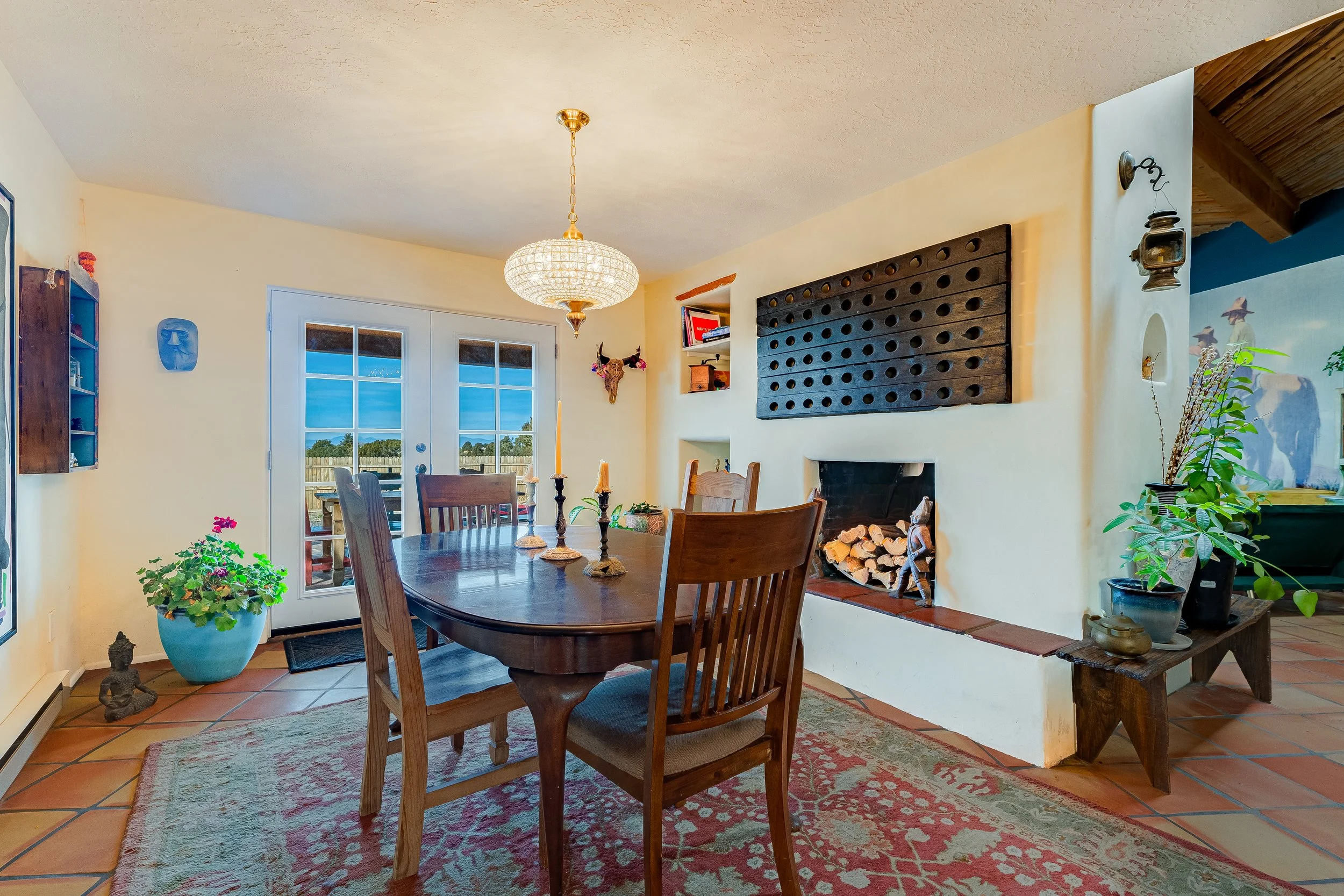Dining room with a wooden table, candles, a fireplace with firewood, potted plants, glass doors, art pieces, and a chandelier.