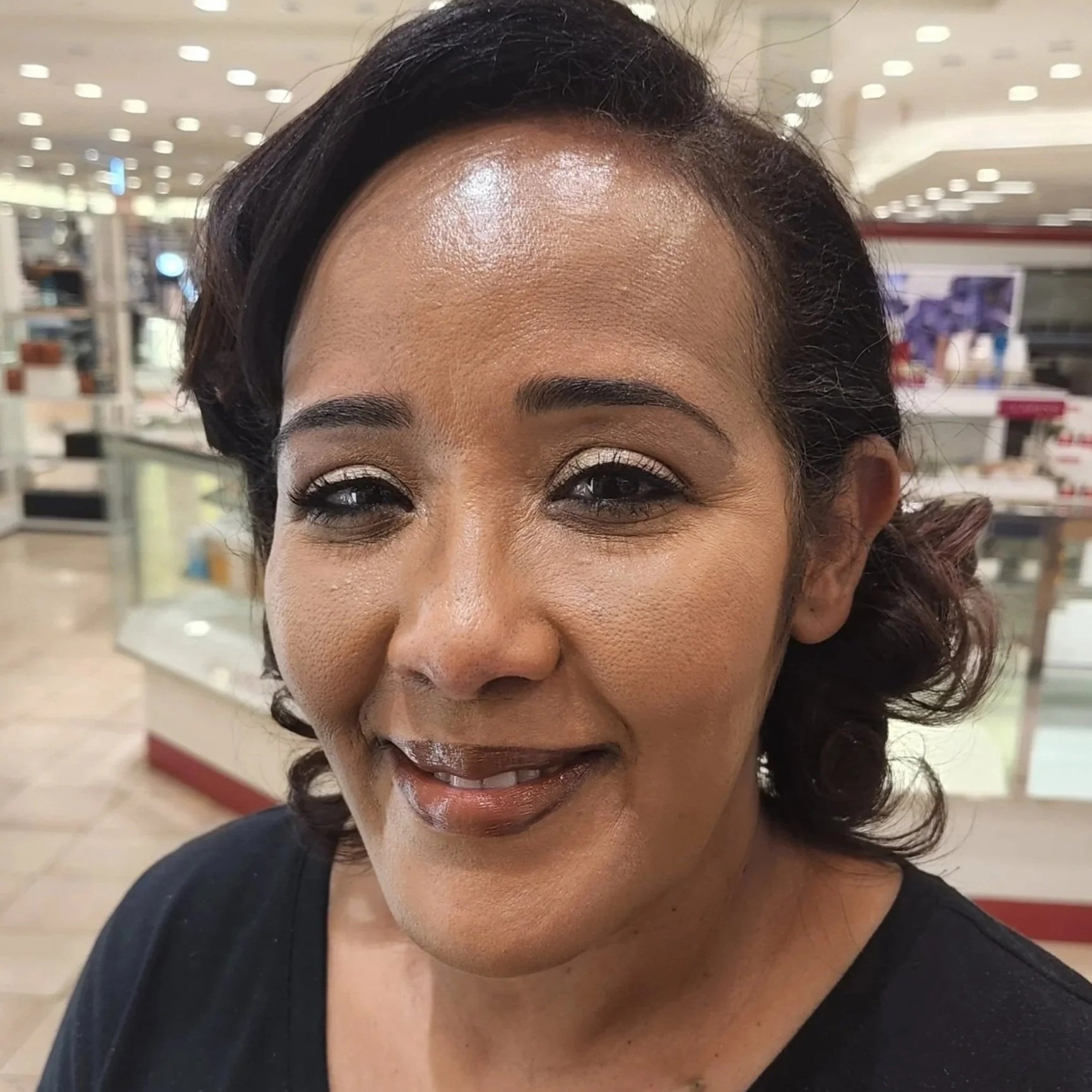 Close-up of a smiling woman with short dark hair and makeup, in a retail store with display shelves and bright ceiling lights in the background.