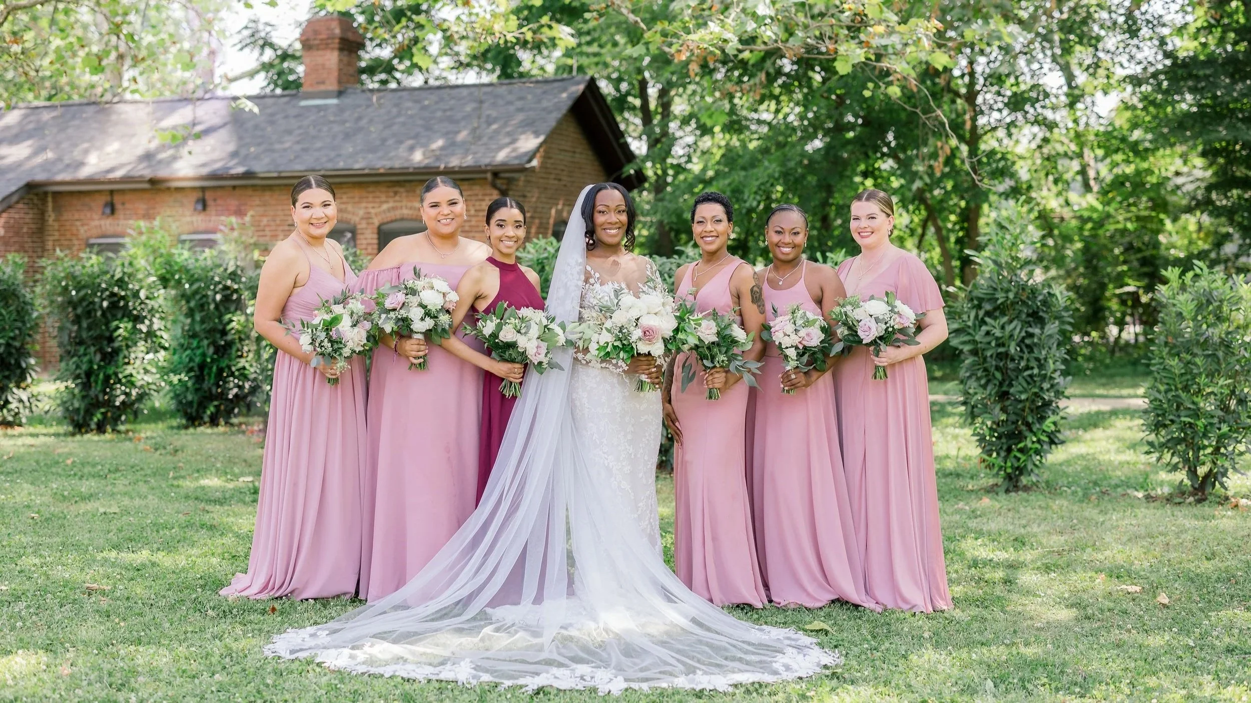 A group of women, including a bride in a white wedding dress and veil, and six bridesmaids in matching pink dresses, standing outdoors with greenery and a brick house in the background, all holding bouquets of flowers and smiling.