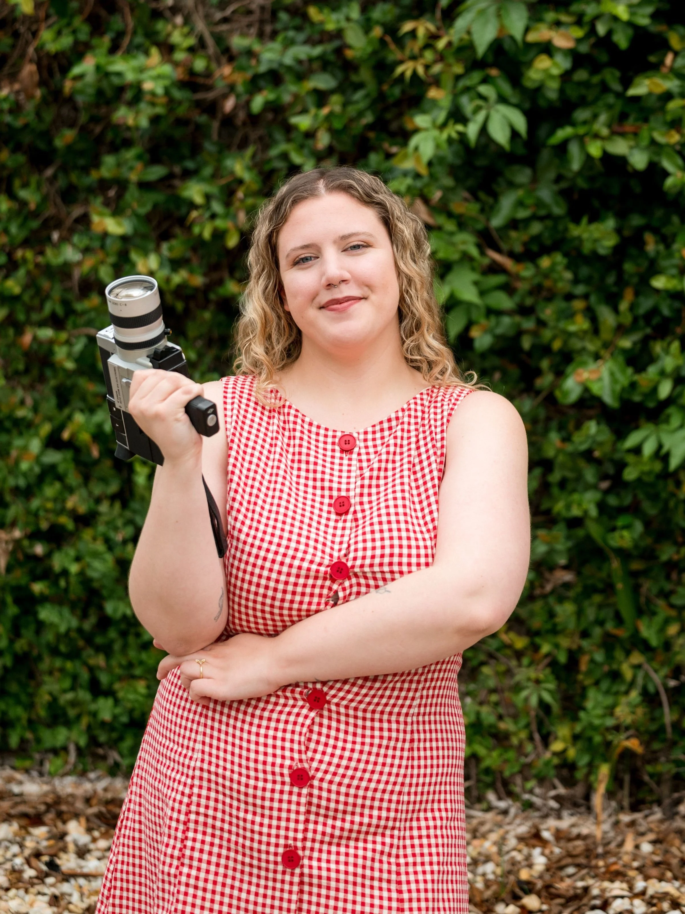 A young woman with curly blonde hair and light skin standing outdoors in front of green shrubbery, wearing a red and white checkered sleeveless dress with red buttons, holding a vintage film camera with a flash attachment.
