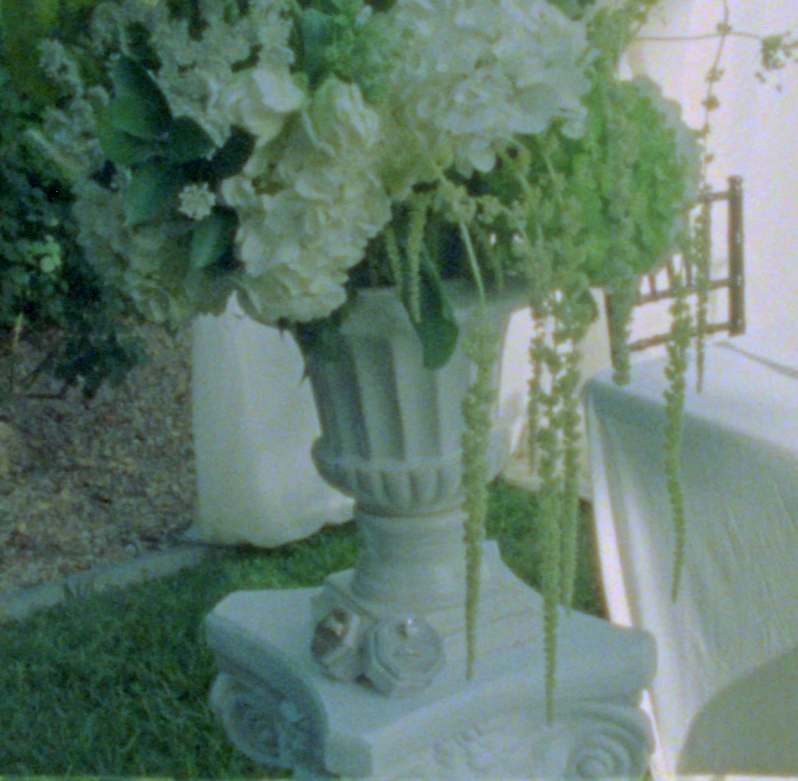 A vase filled with white and light green flowers, sitting on a stone pedestal, with greenery and outdoor furniture in the background.