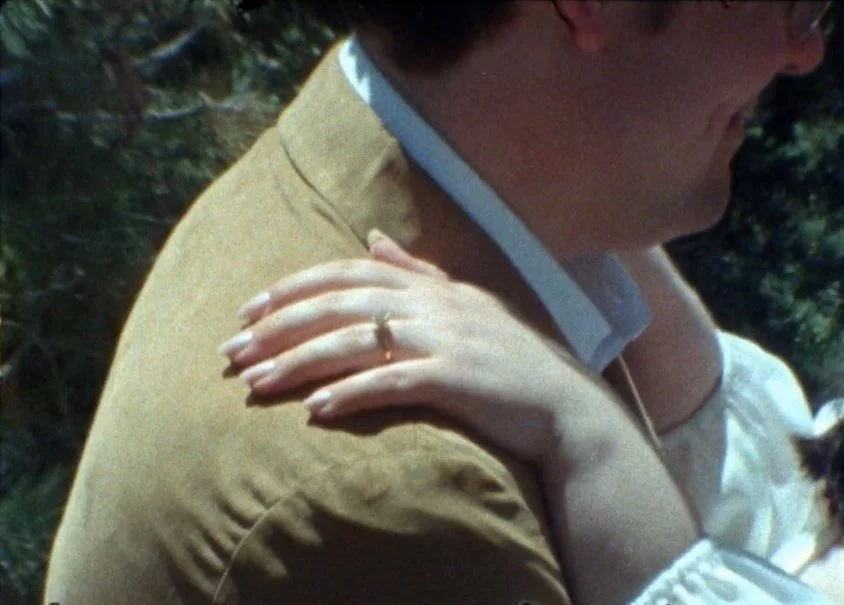 Close-up of a man in a light brown jacket and a woman with her hand resting on his shoulder, with a visible wedding ring on her finger.
