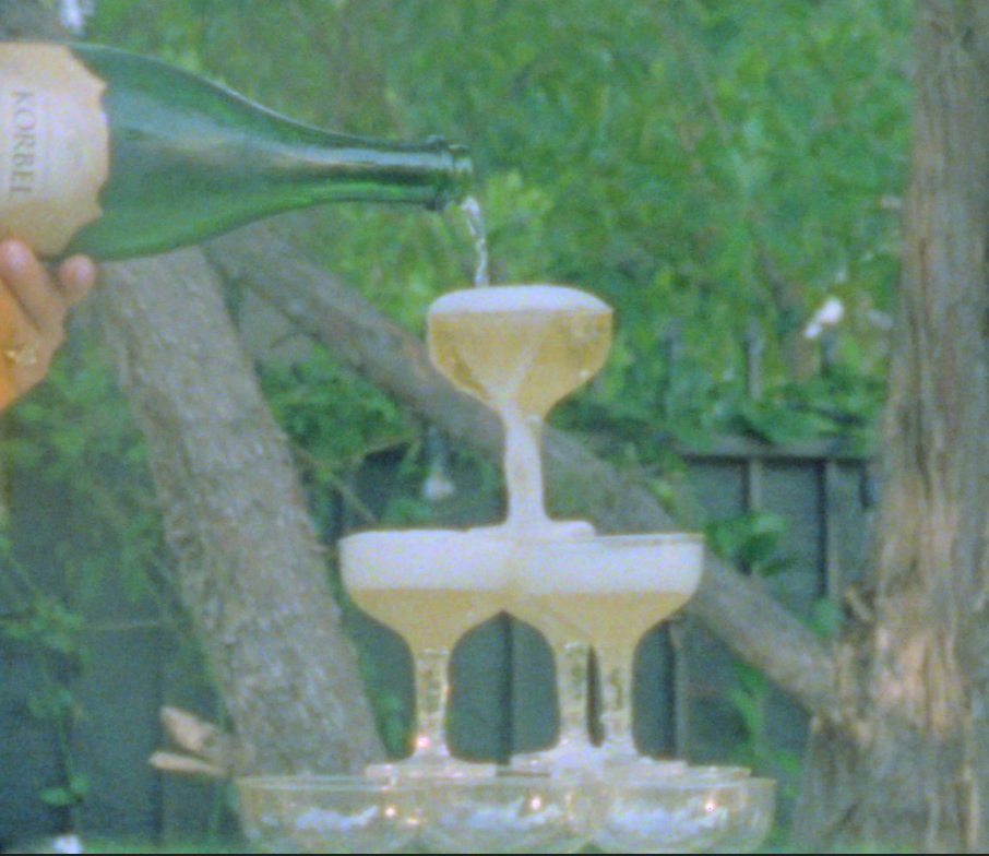 A person is pouring champagne into a tiered pyramid of champagne glasses outdoors, surrounded by trees.