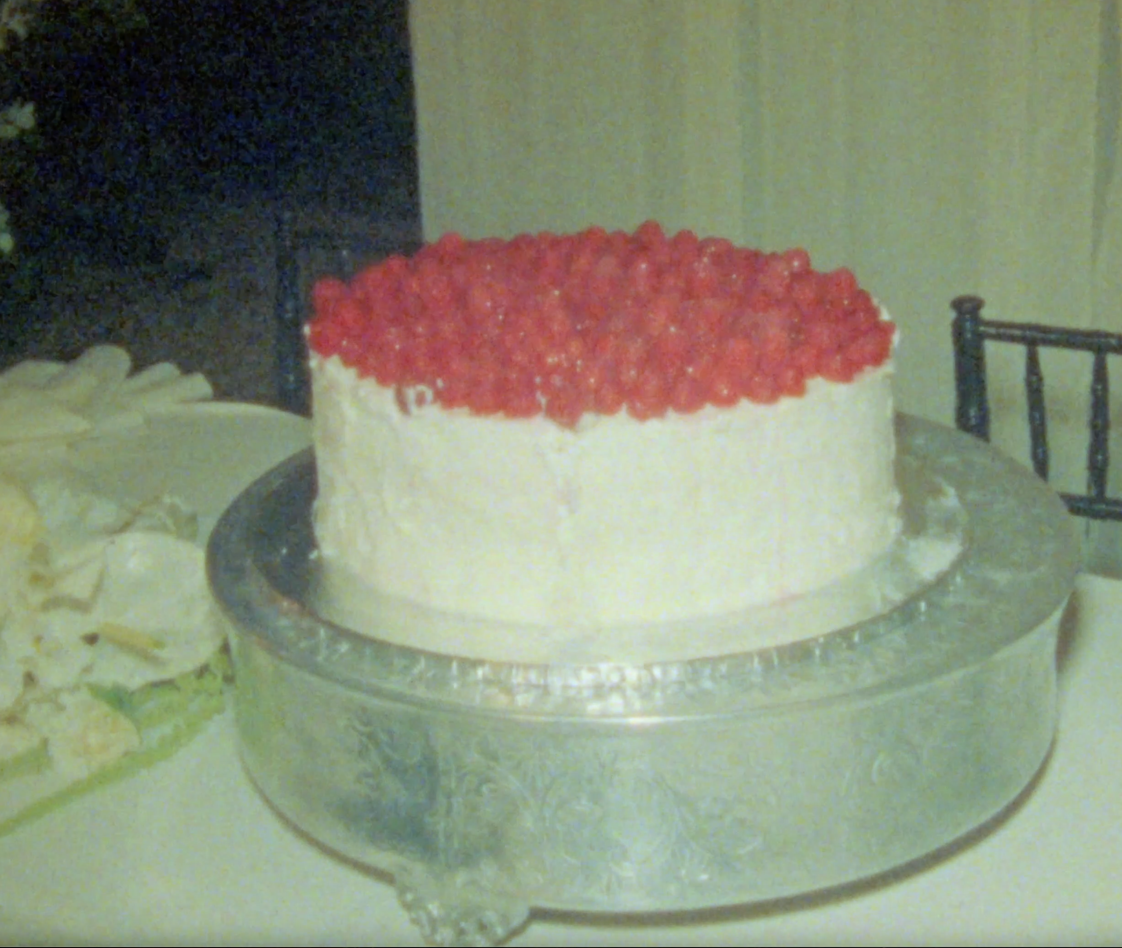 A round cake with white frosting topped with red crushed berries, placed on a silver cake stand.