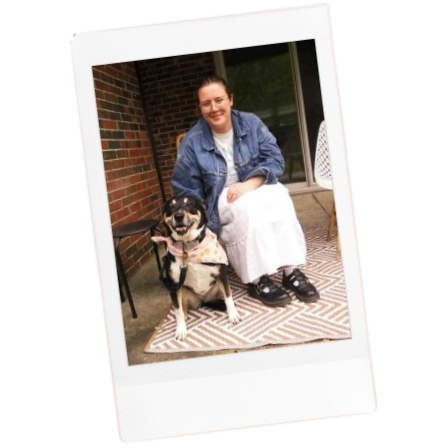 A woman sitting outdoors on a porch next to a black and tan dog wearing a pink bandana, with a brick wall and a sliding glass door in the background.