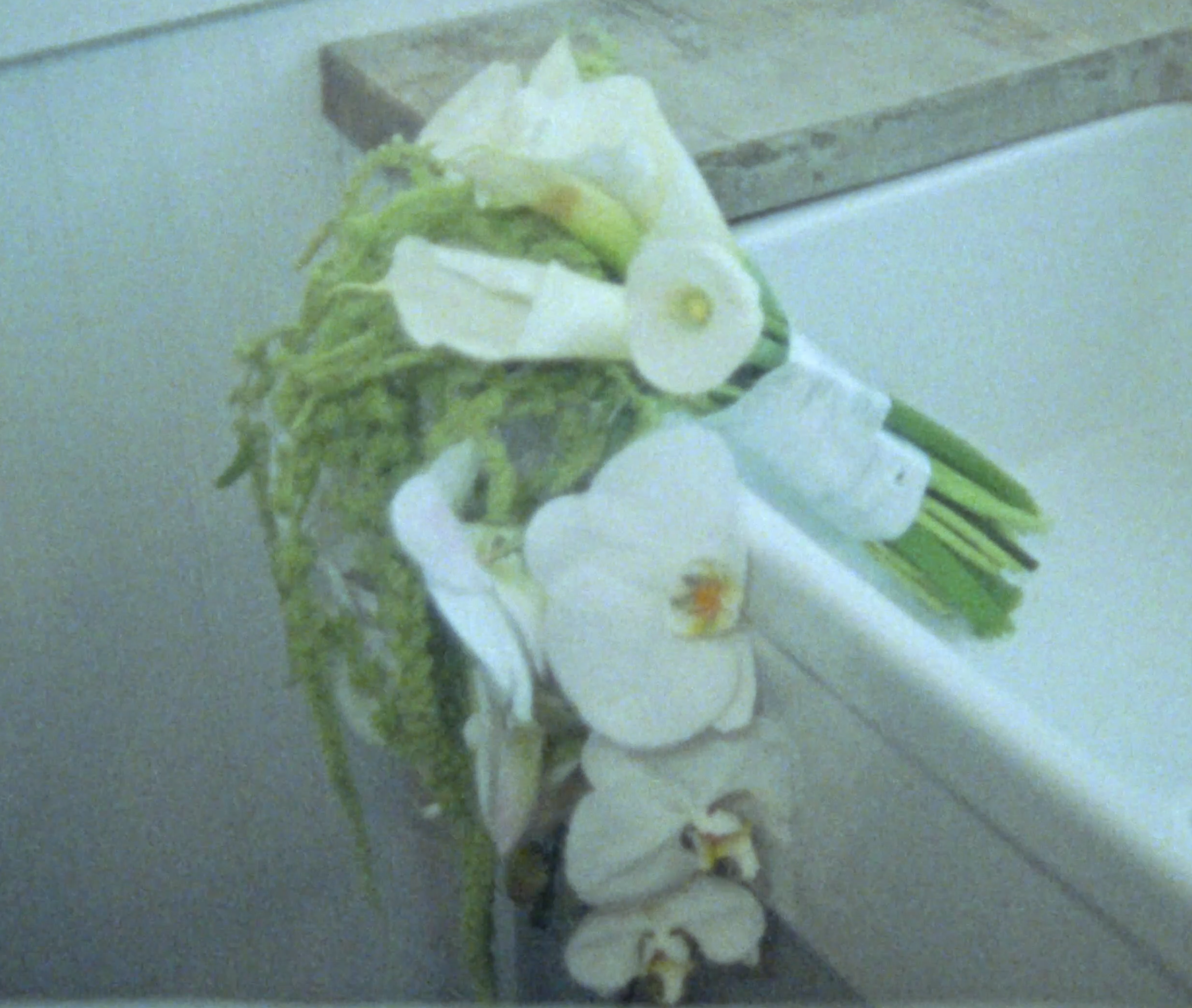 A bunch of white orchids with green stems resting on a white surface next to a wooden piece, possibly a table or shelf.