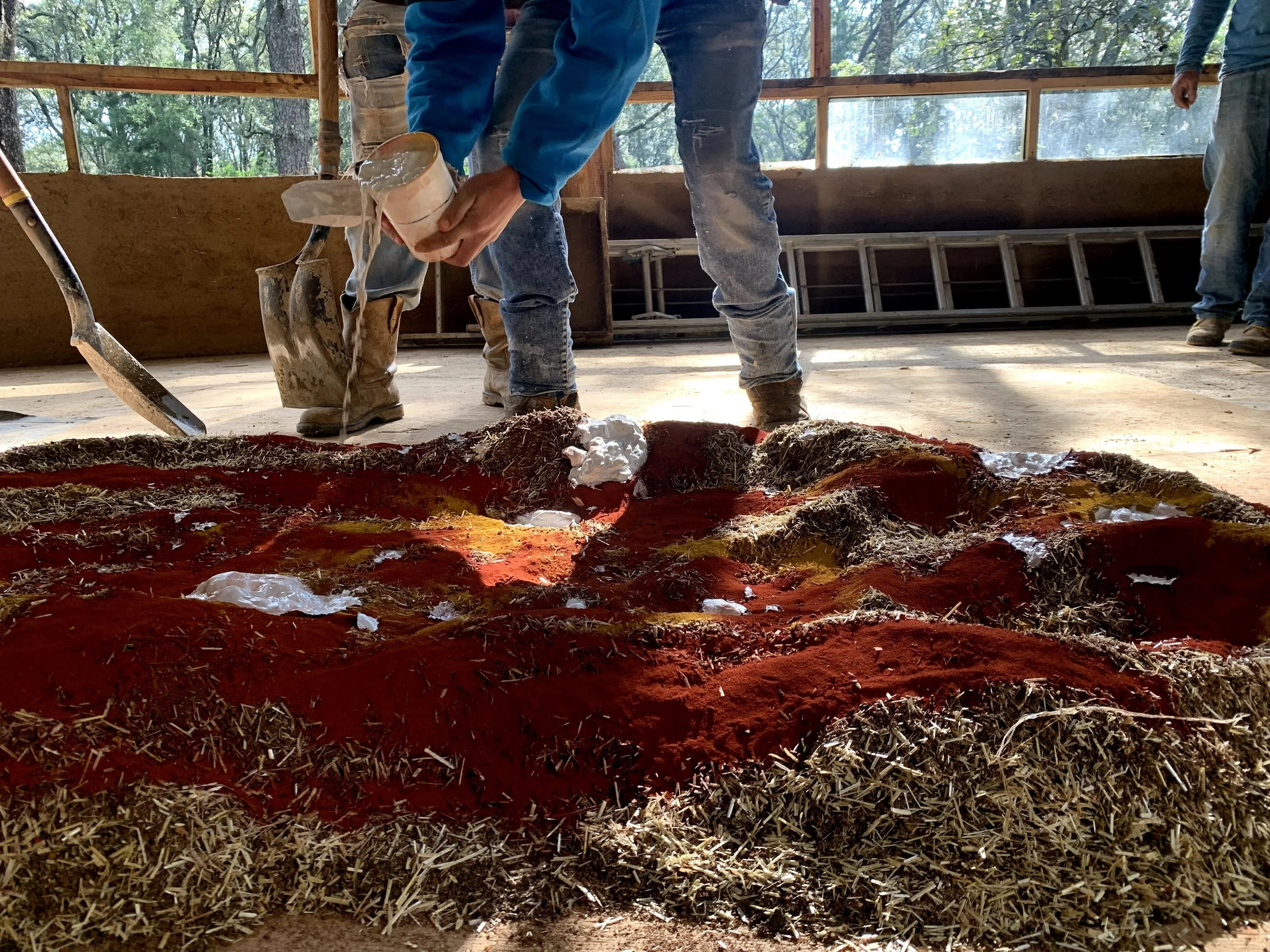 Persona participando de la bioconstrucción, vertiendo líquido sobre mezcla de tierra y hierbas como parte de una práctica de arquitectura sustentable