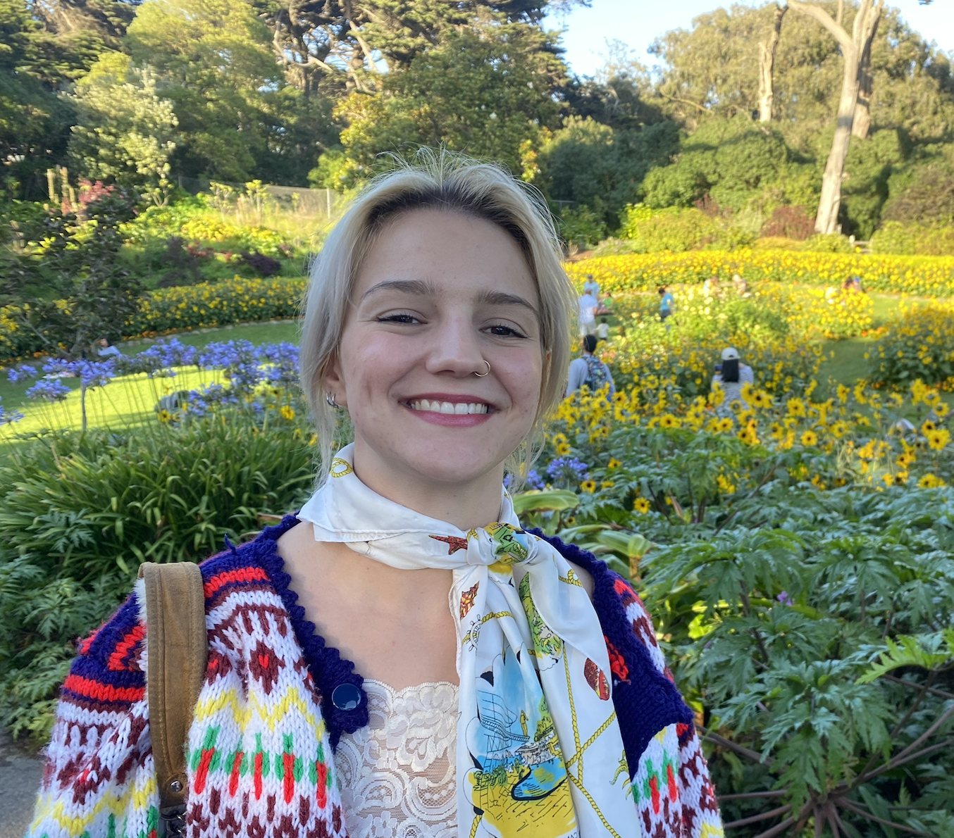 A young woman with blonde hair and fair skin smiling outdoors in a garden filled with colorful flowers, including purple and yellow blooms, during daytime.