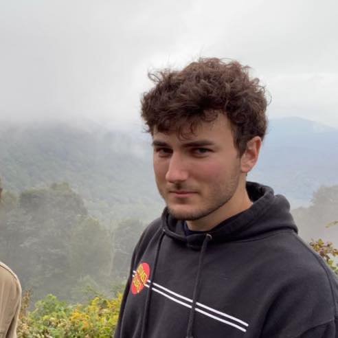 Young man with curly brown hair standing outdoors with a foggy mountain landscape in the background.