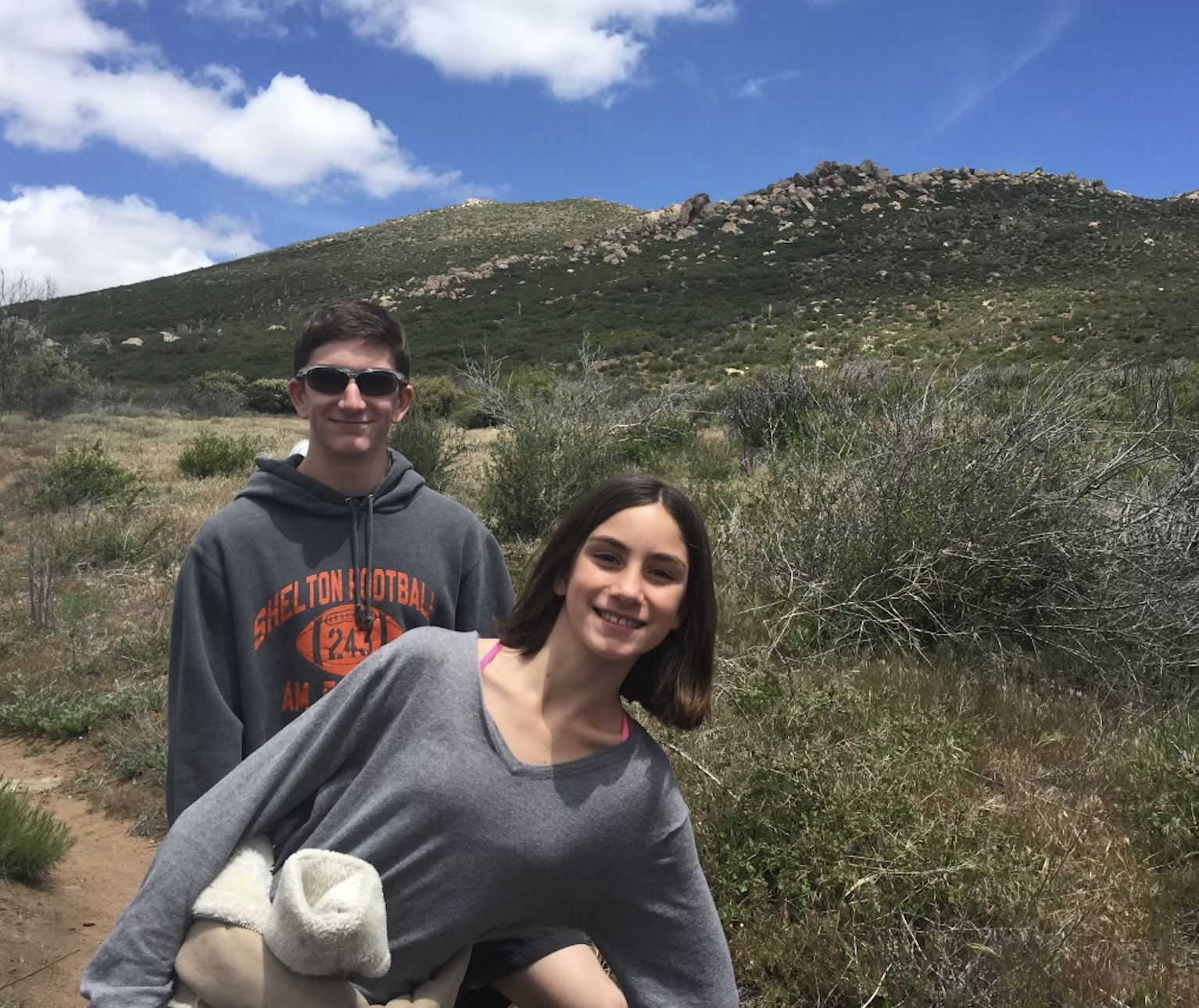 A young man and woman hiking outdoors in a mountainous landscape with a bright blue sky and a few clouds.