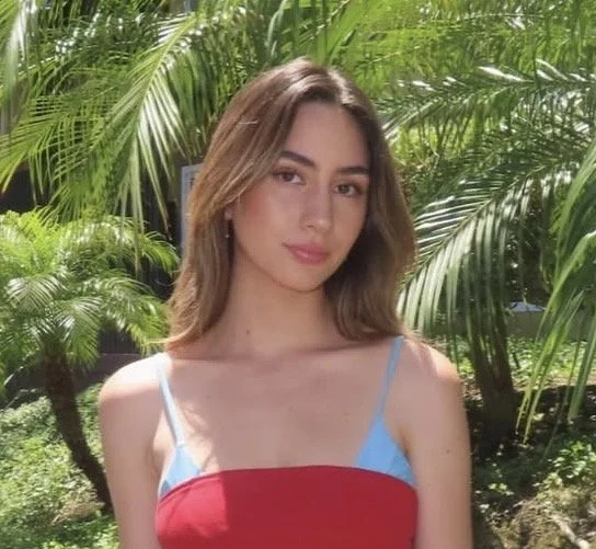 Young woman with shoulder-length brown hair wearing a colorful tank top, standing outdoors with lush green tropical plants in the background.