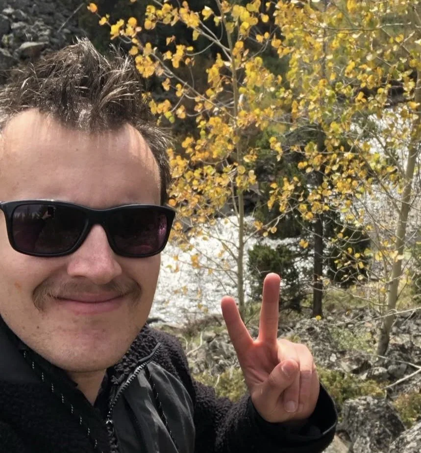 A man with sunglasses and a mustache taking a selfie outdoors, showing a peace sign with his hand, with yellow autumn leaves and rocks in the background.