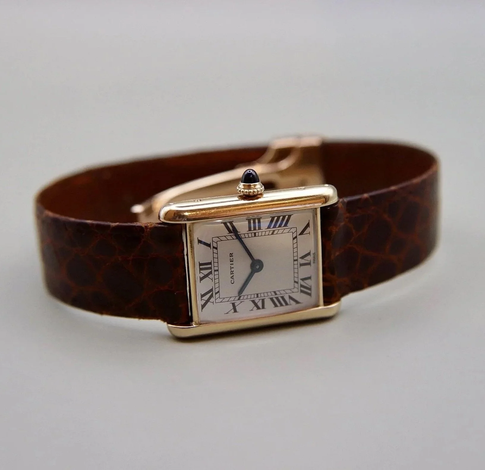 A gold Cartier watch with a square face, Roman numerals, and a black cabochon on the crown, attached to a brown leather strap.