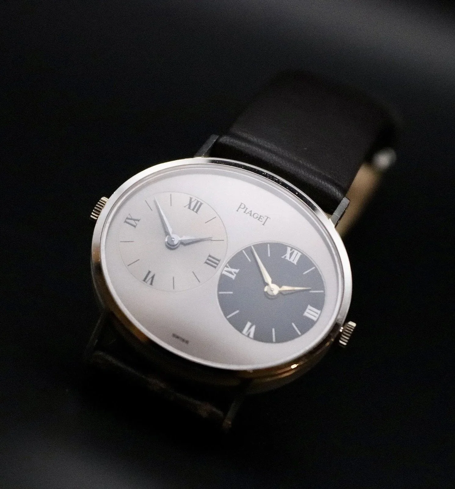 A luxury wristwatch with a black leather strap, featuring two dials with roman numerals, one in silver and the other in dark gray, on a dark surface.