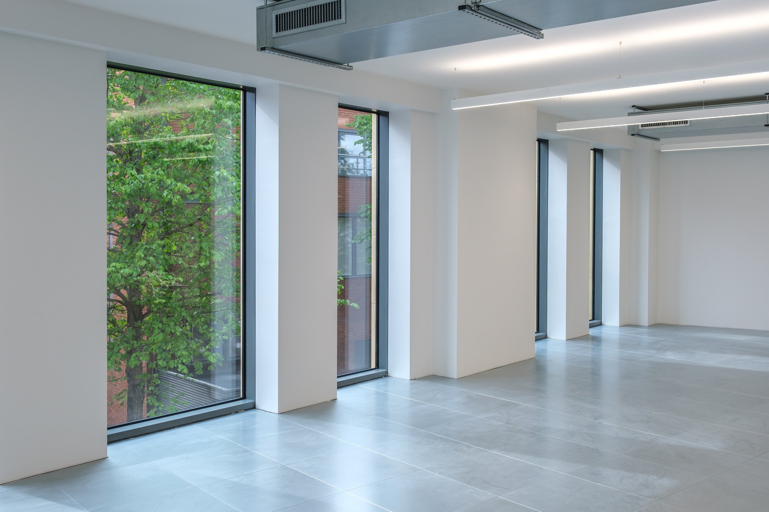 Empty modern office space with large windows, gray tiled floor, and overhead lighting fixtures; trees visible outside.