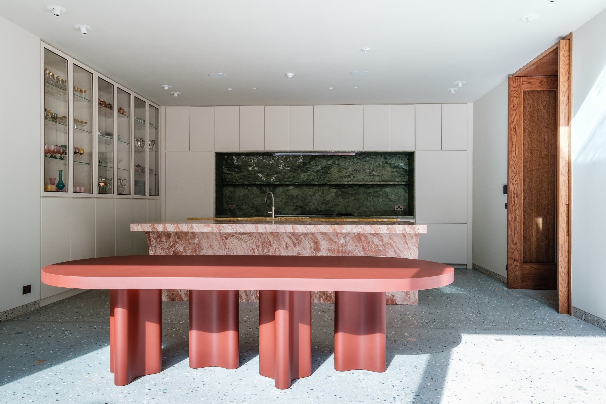 Modern kitchen interior with a large red table, marble island, and glass cabinet filled with glassware.