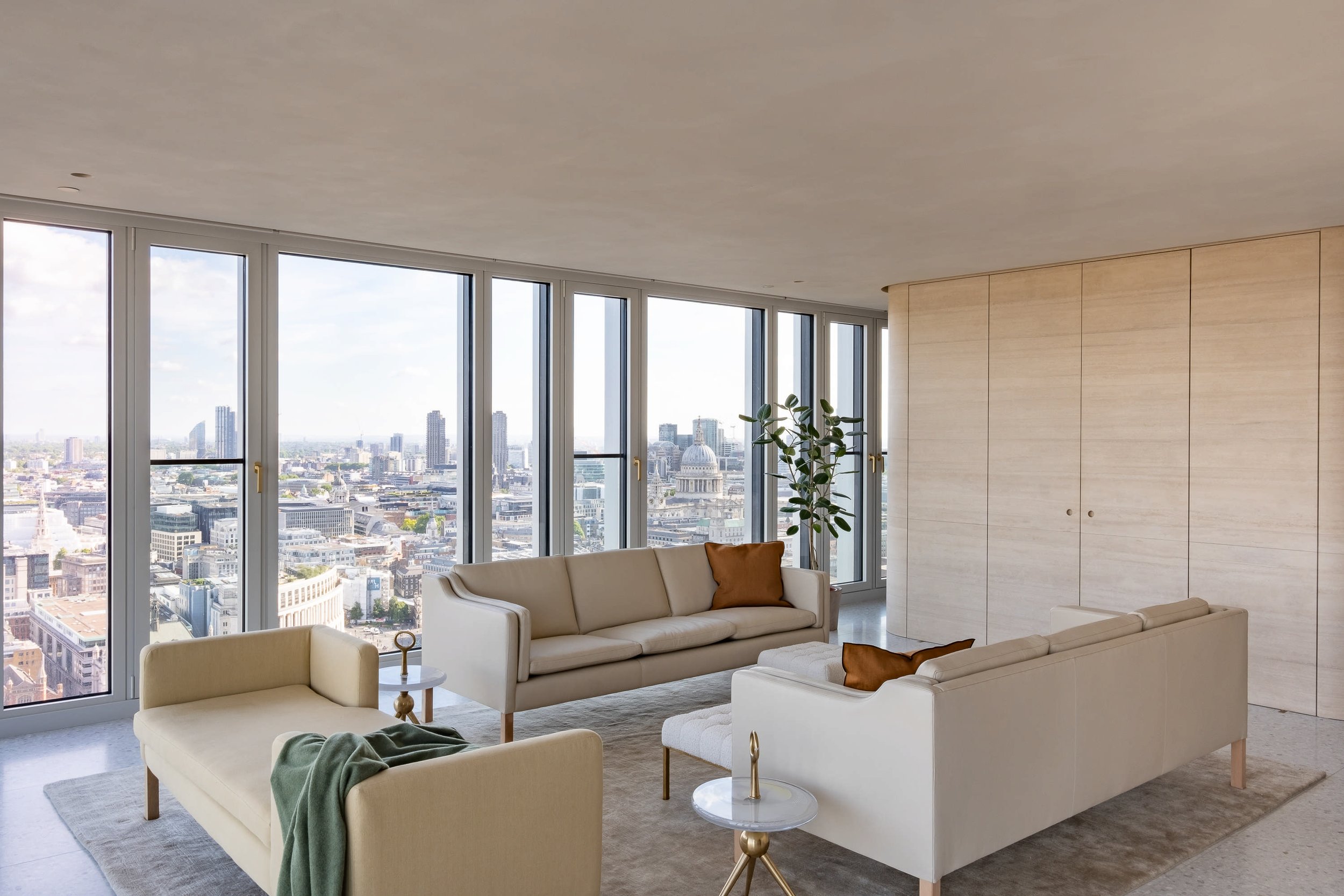 Modern living room with large windows showcasing a city skyline, beige sofas with brown cushions, a green potted plant, and light-colored wooden wall panels.