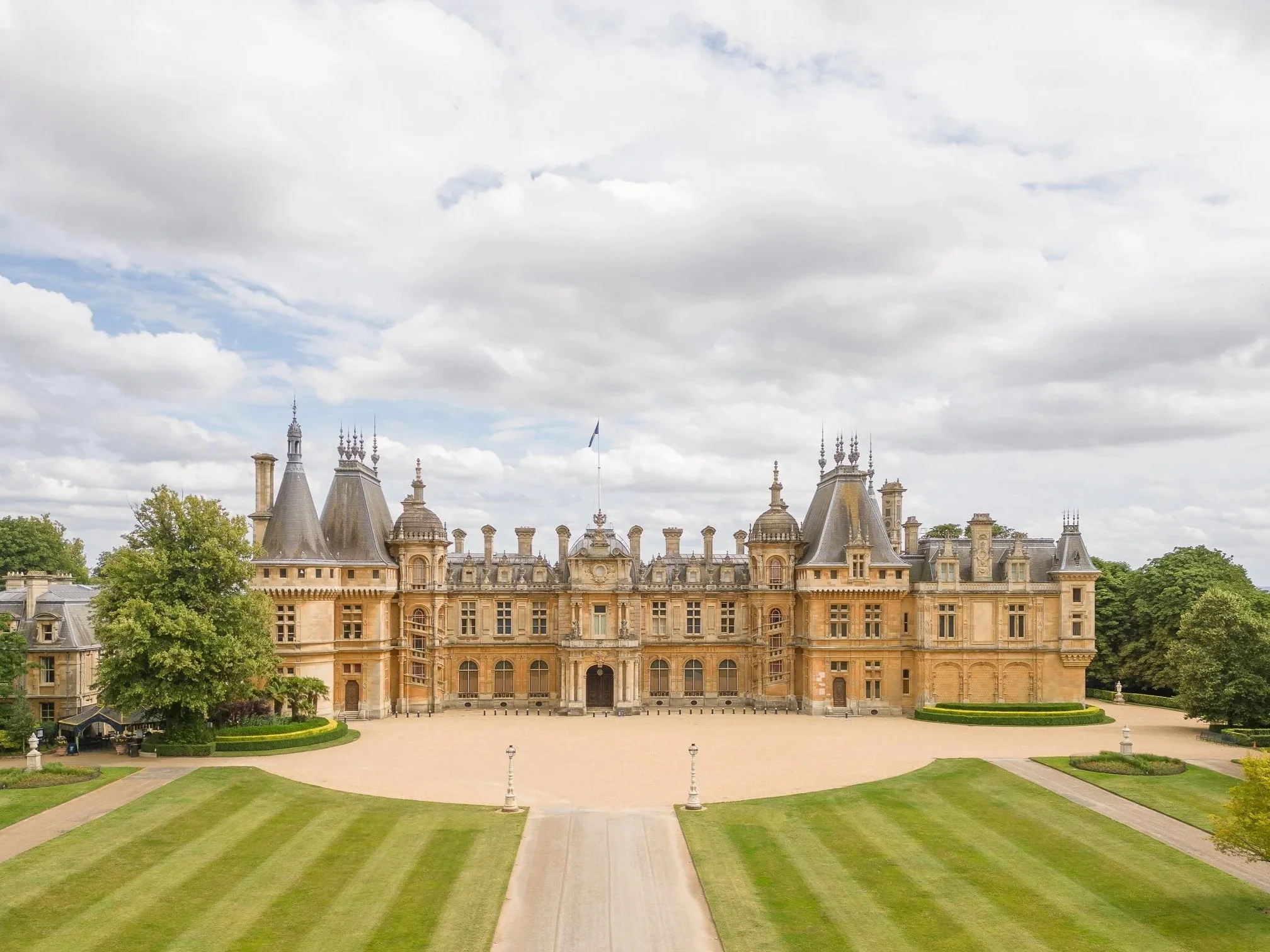 A large historic castle with multiple towers and turrets, surrounded by well-maintained gardens and green lawns under a cloudy sky.