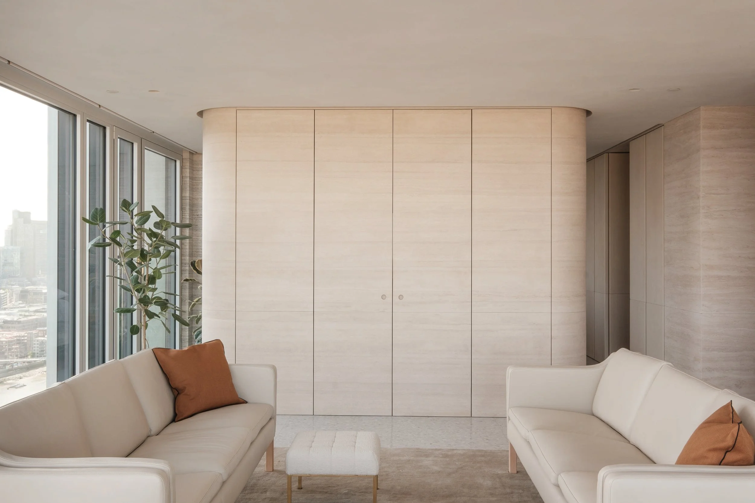 Modern living room with two white sofas, one with a brown pillow, a small beige ottoman, tall windows with city view, and a built-in wooden cabinet wall.