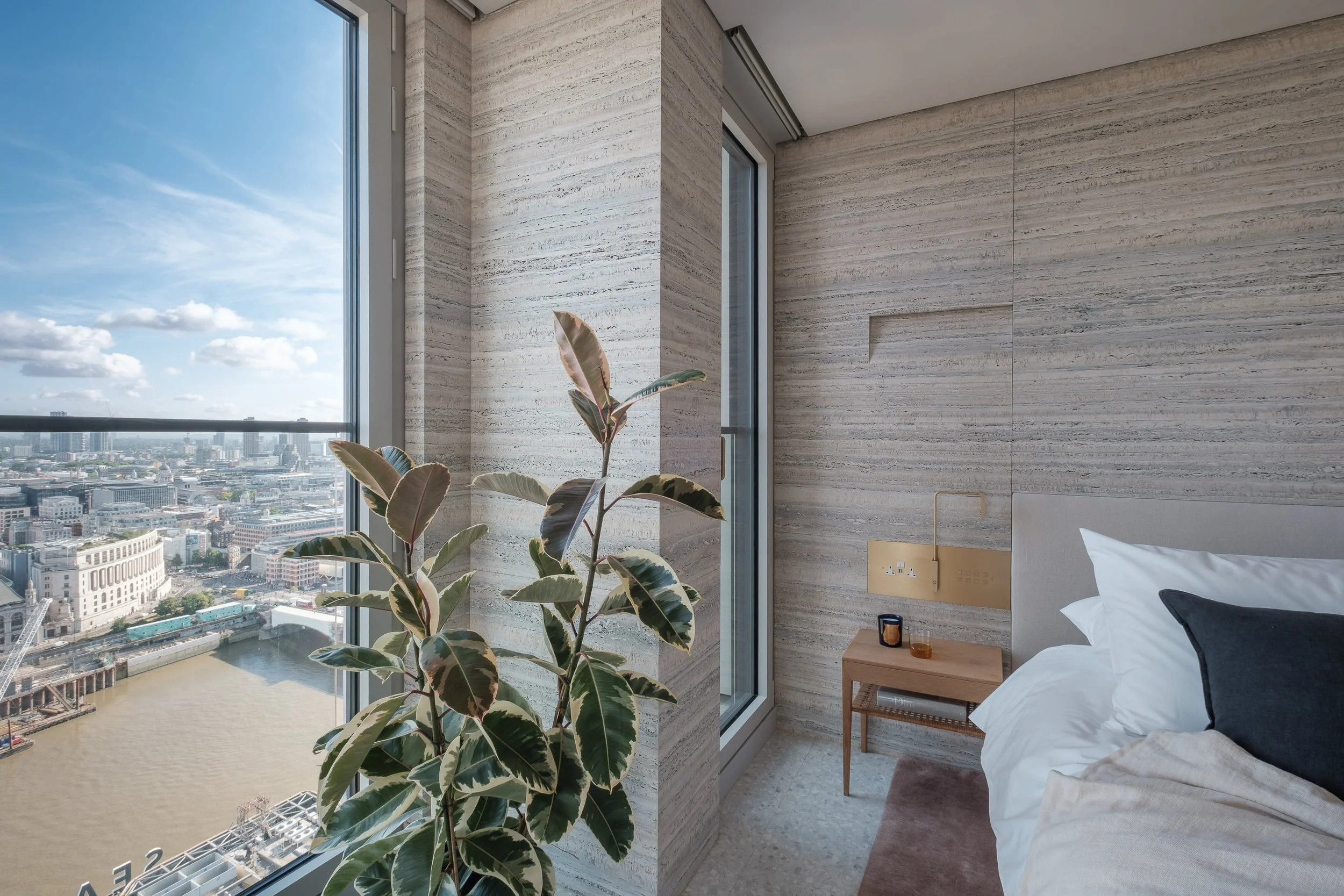 A modern bedroom with a large window overlooking and river. The room features beige marble walls, a bed with white linens and a black pillow, and a wooden bedside table with decorative items. A tall, broad-leafed plant is near the window, and natural