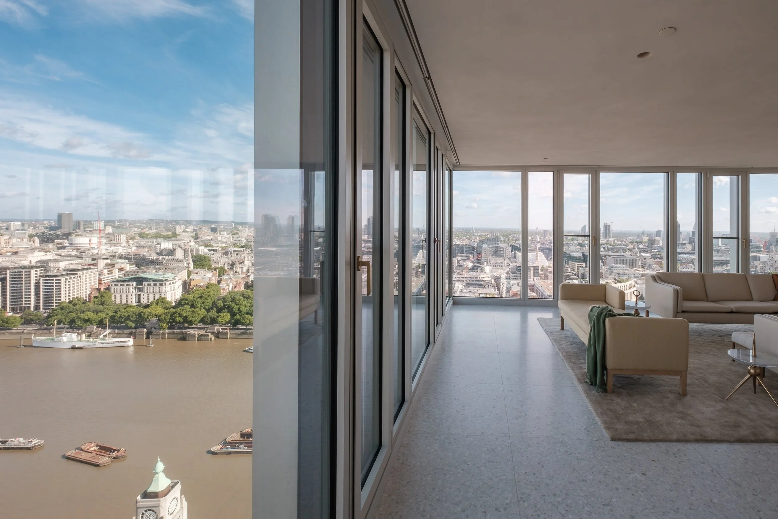 Luxury apartment living room with large windows offering a city of London view, beige sofas, a glass coffee table, and a rug.
