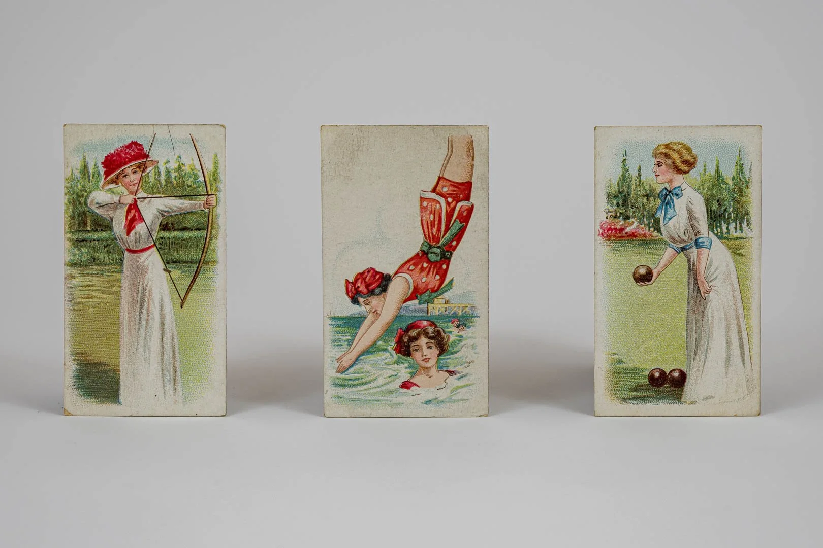 Three vintage playing cards, each featuring a woman engaged in outdoor sports: archery, swimming, and lawn bowling.