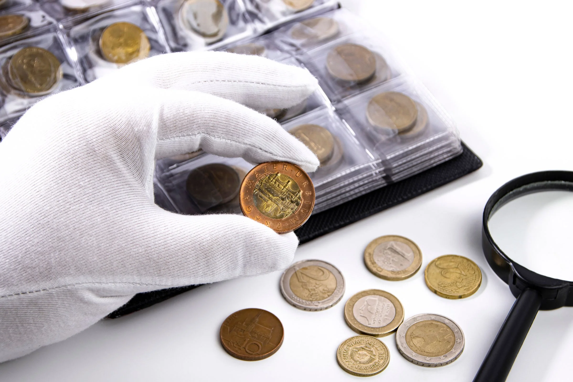 A gloved hand holding a collectable coin taken from an album along with a magnifying glass on a white surface