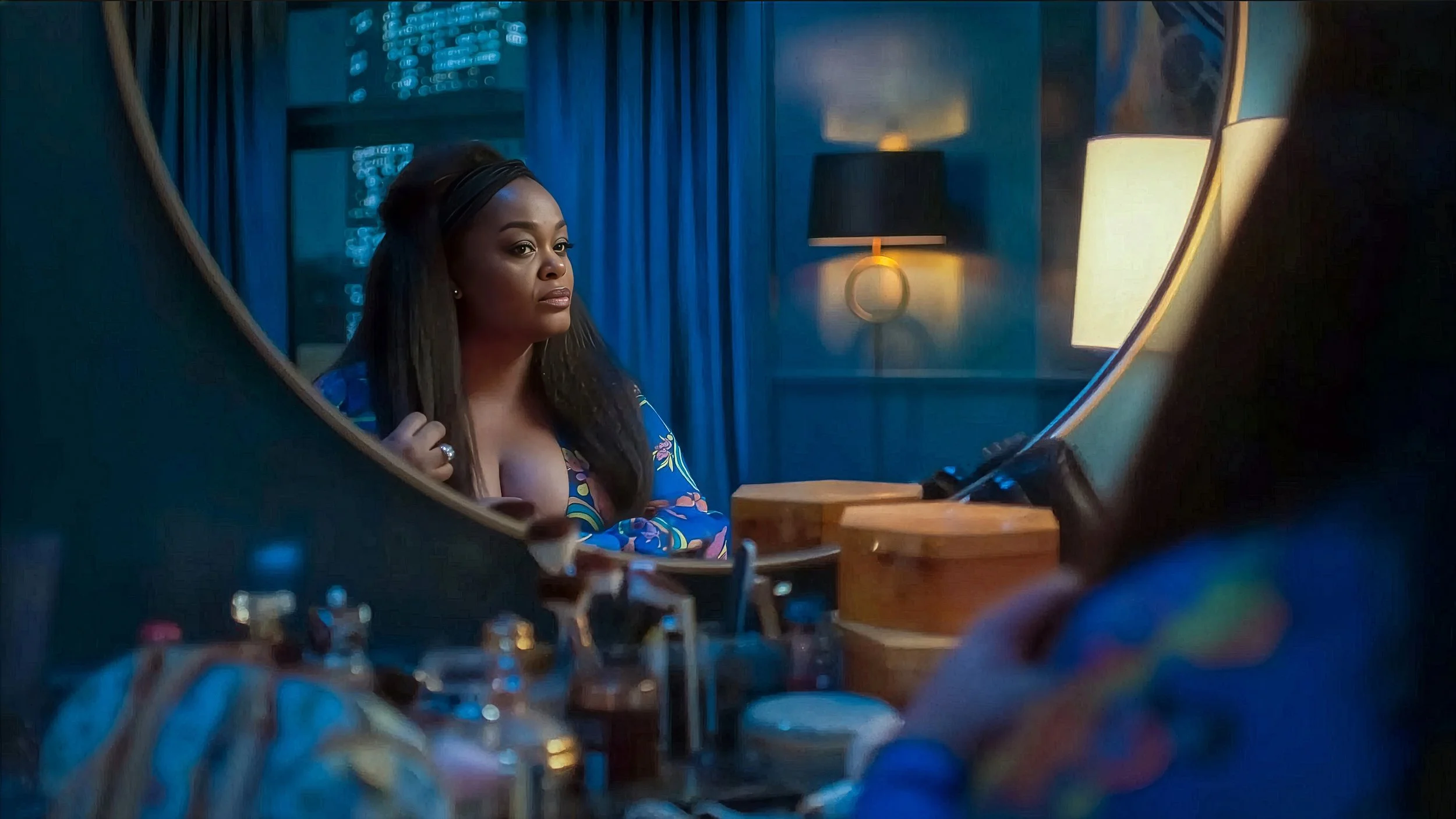 A woman with long dark hair and a colorful patterned dress looking into a mirror in a dimly lit room with warm yellow lamps and blue curtains.