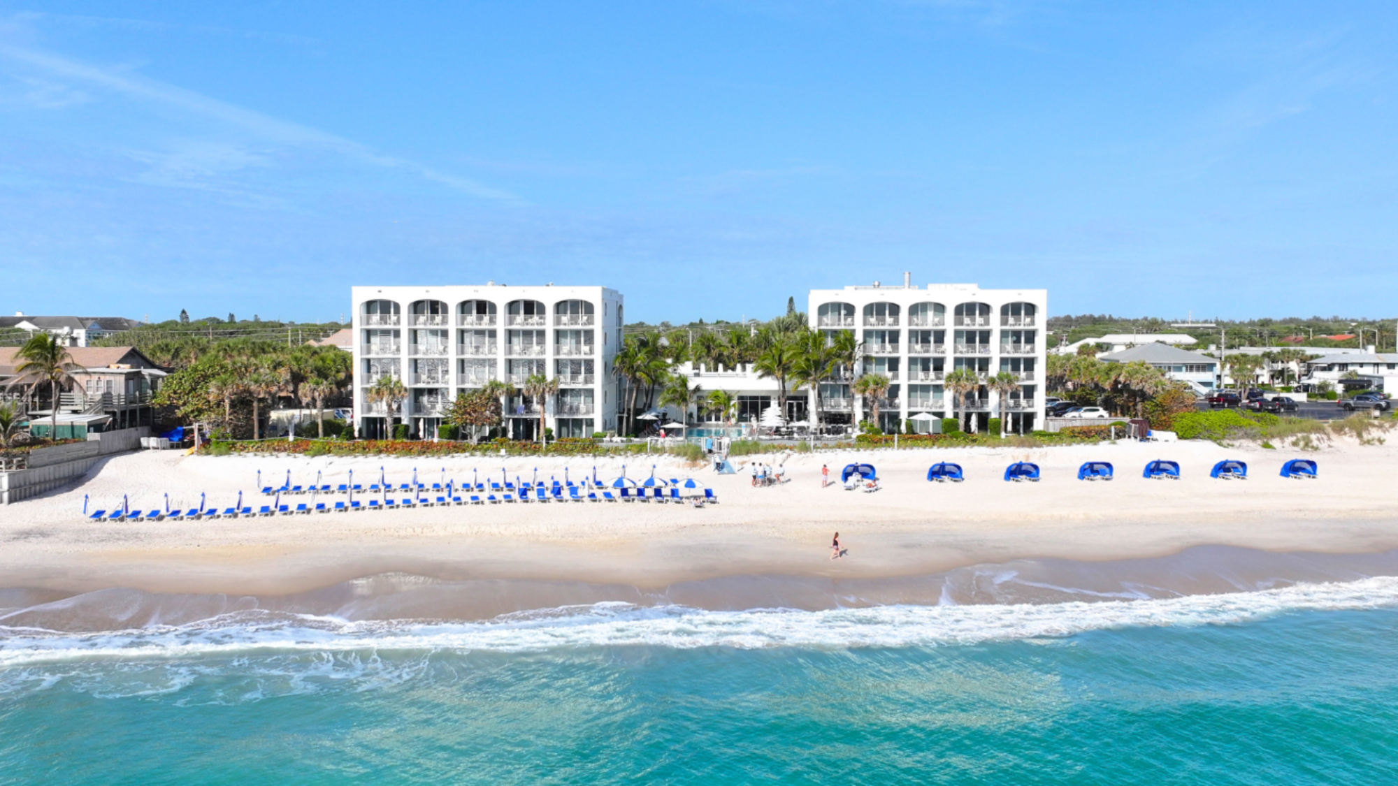Beachfront resort with white buildings, beach chairs and umbrellas, palm trees, and ocean waves.