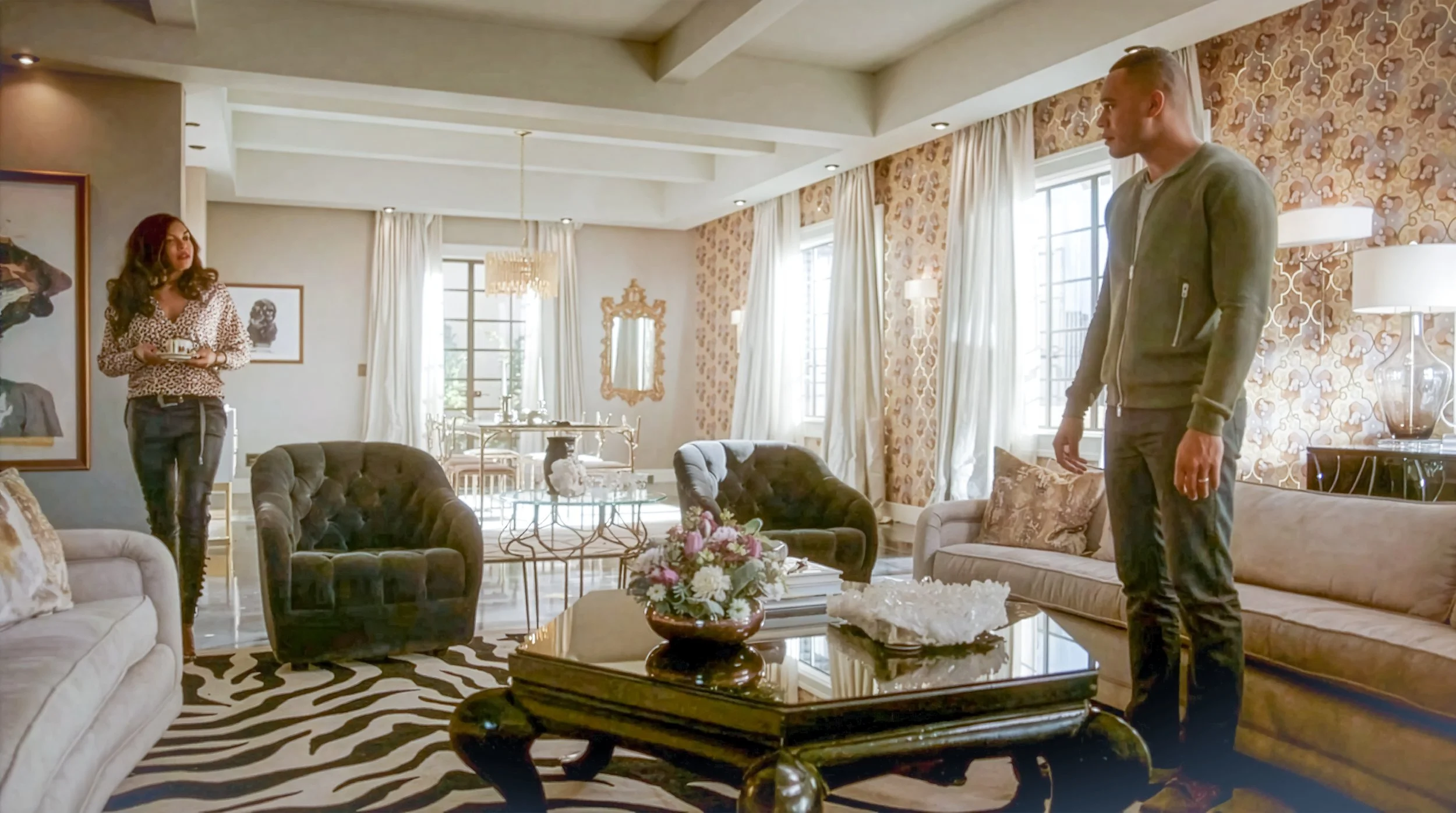 A woman with red hair and a leopard print blouse holding a tray stands in a well-decorated living room facing a man with short hair and a gray jacket. The room has large windows, white curtains, a zebra print rug, and various chairs and a coffee table with a floral arrangement.