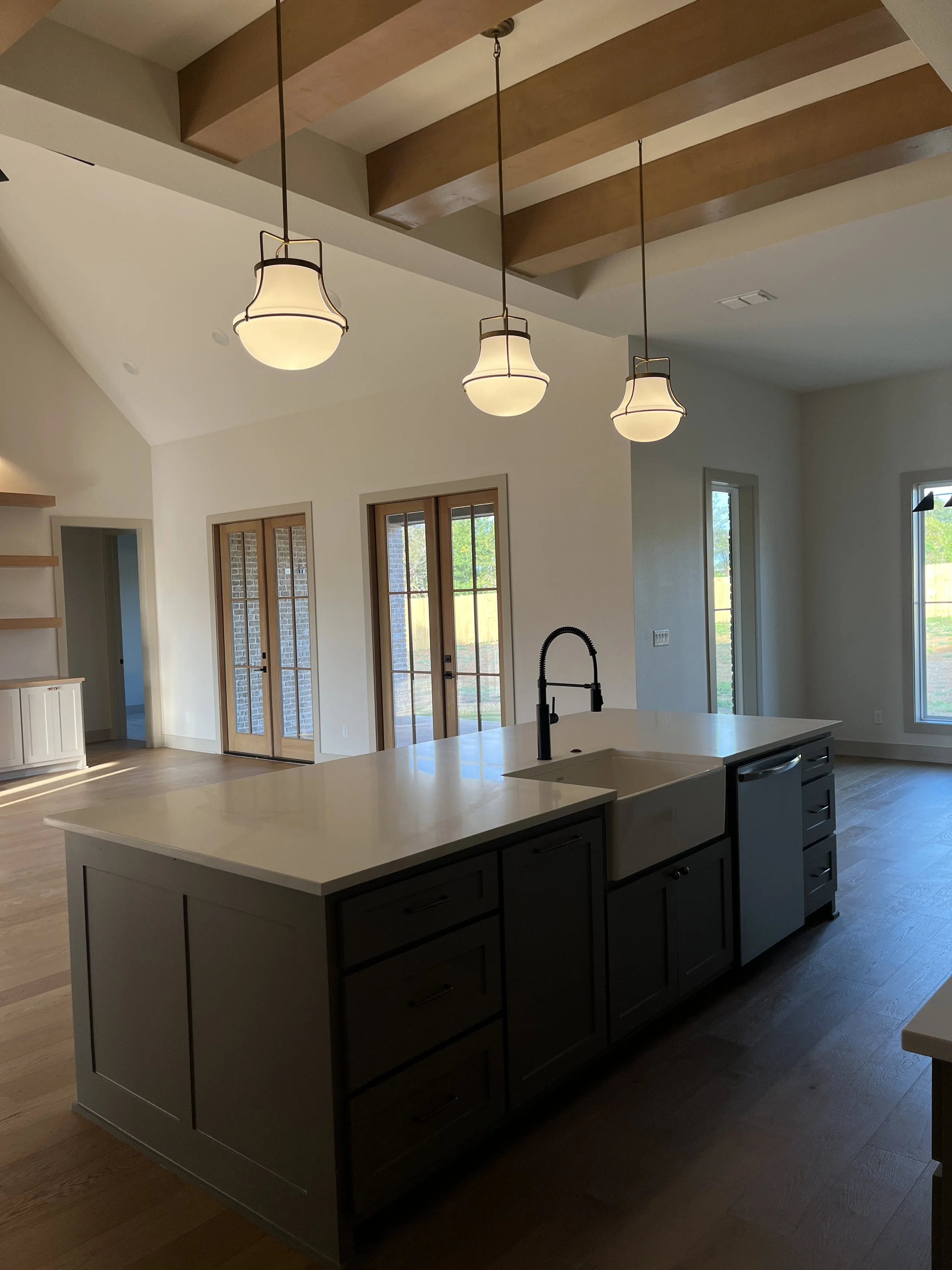Modern kitchen with dark cabinets, white countertops, farmhouse sink, black faucet, pendant lights, wooden ceiling beams, and double glass doors leading outside.
